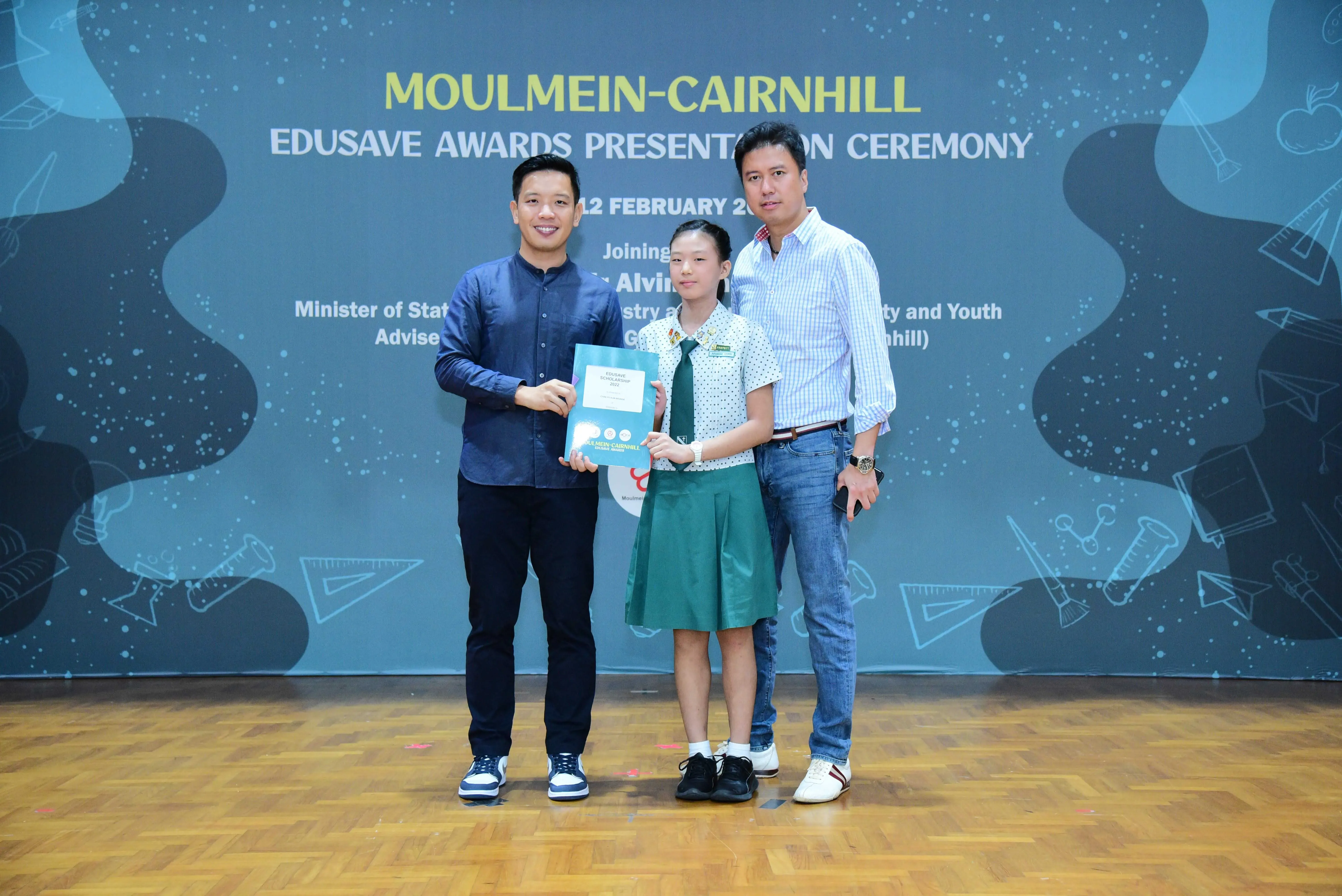 A girl in school uniform holds an award certificate between two men at the Moulmein-Cairnhill Edusave Awards Ceremony.