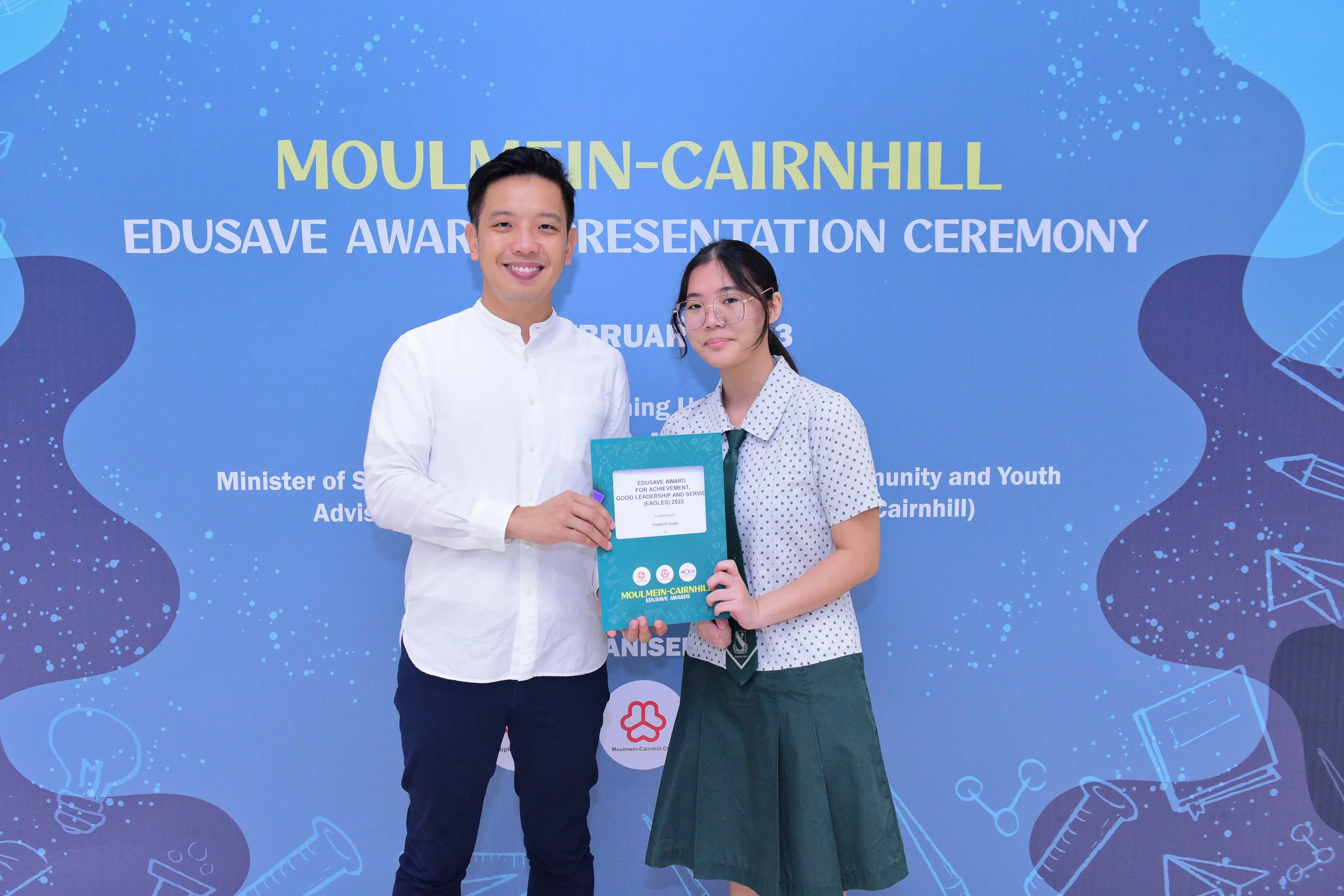 A man and a young female student hold an Edusave award folder at the Moulmein-Cairnhill Award Presentation Ceremony.