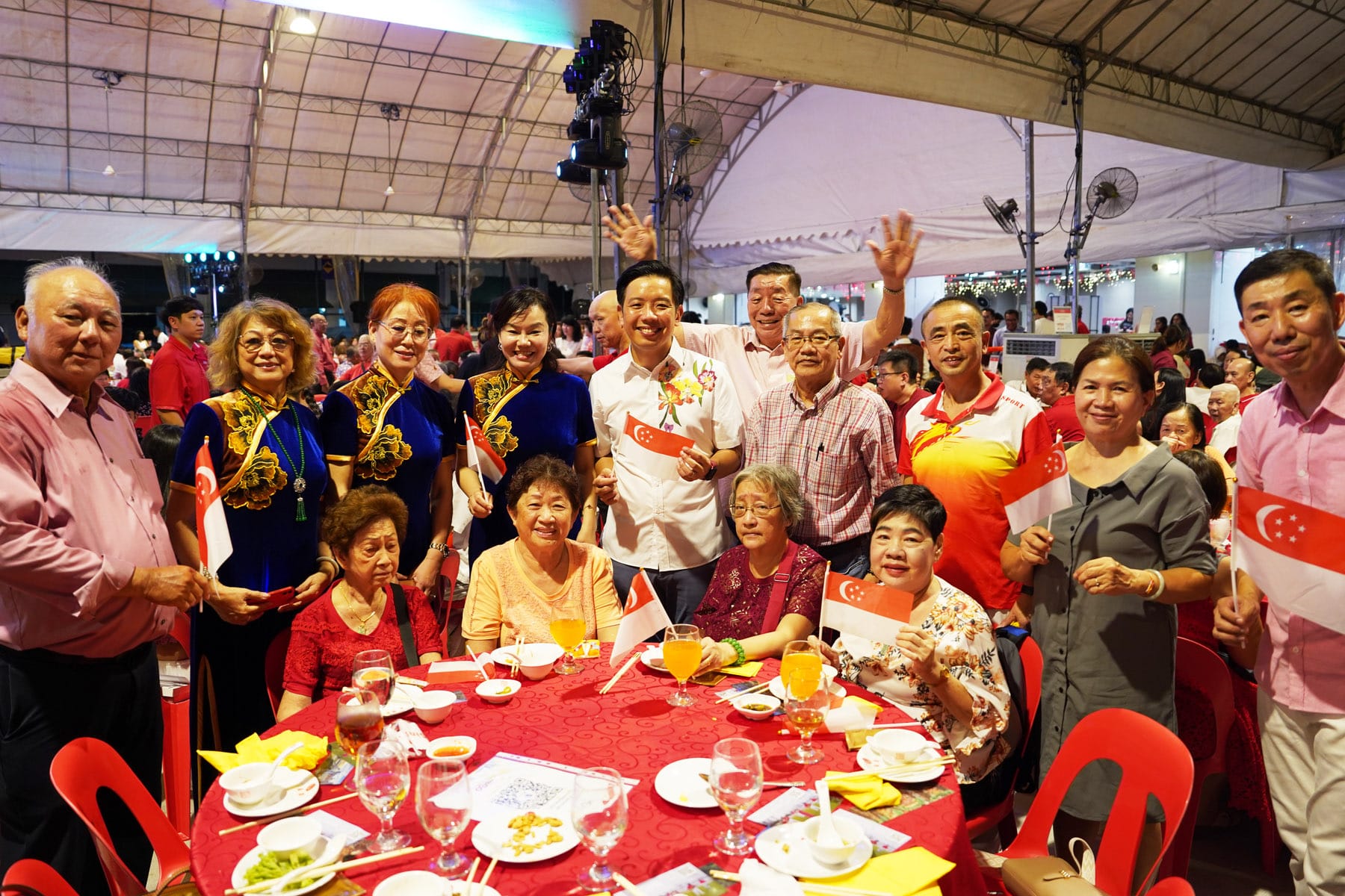 A large group of people pose together at a banquet, holding Singapore flags, seated and standing around a red-clothed table.