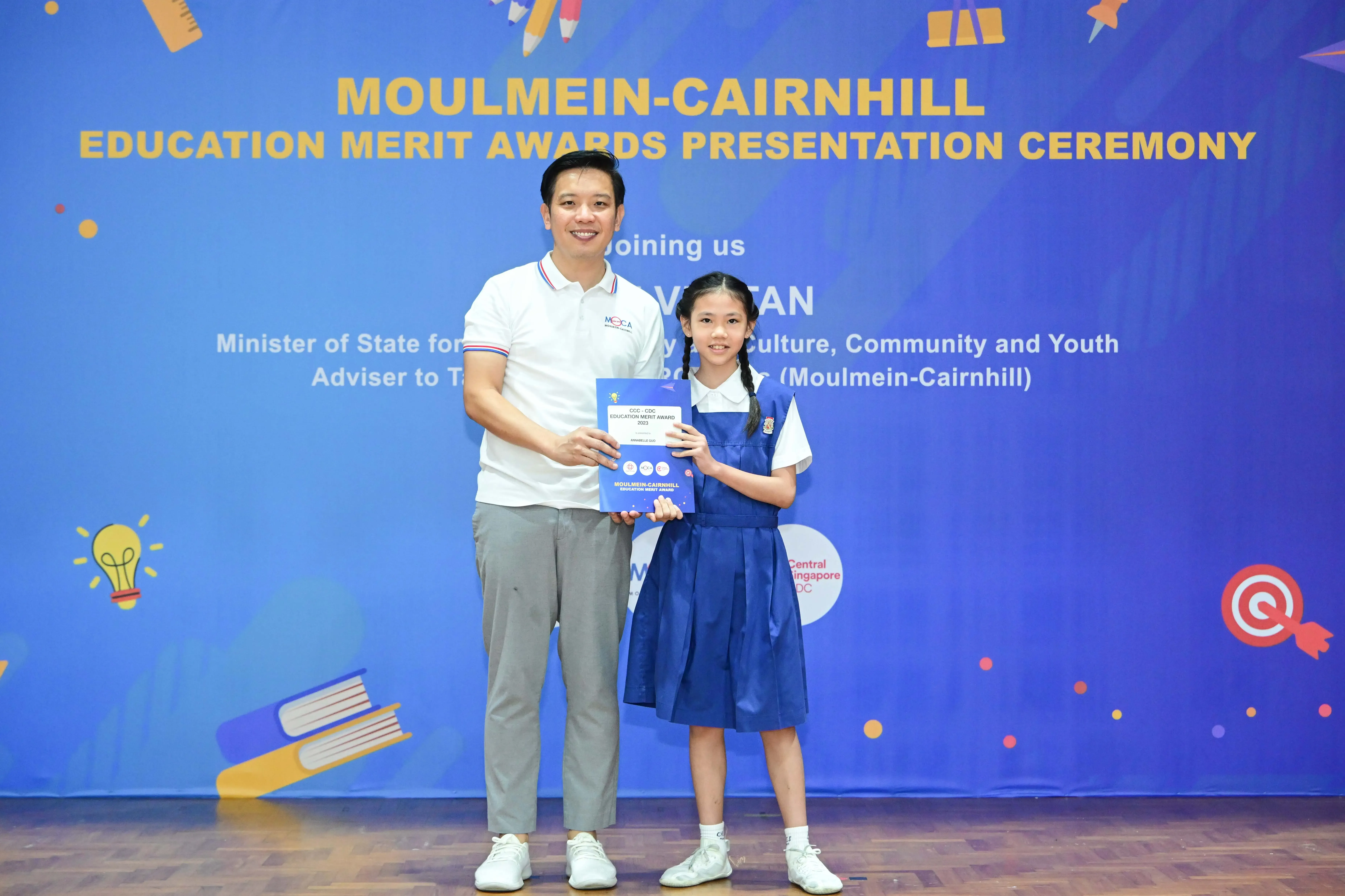 A man and a young girl in school uniform hold an award certificate at the Moulmein-Cairnhill Education Merit Awards ceremony.