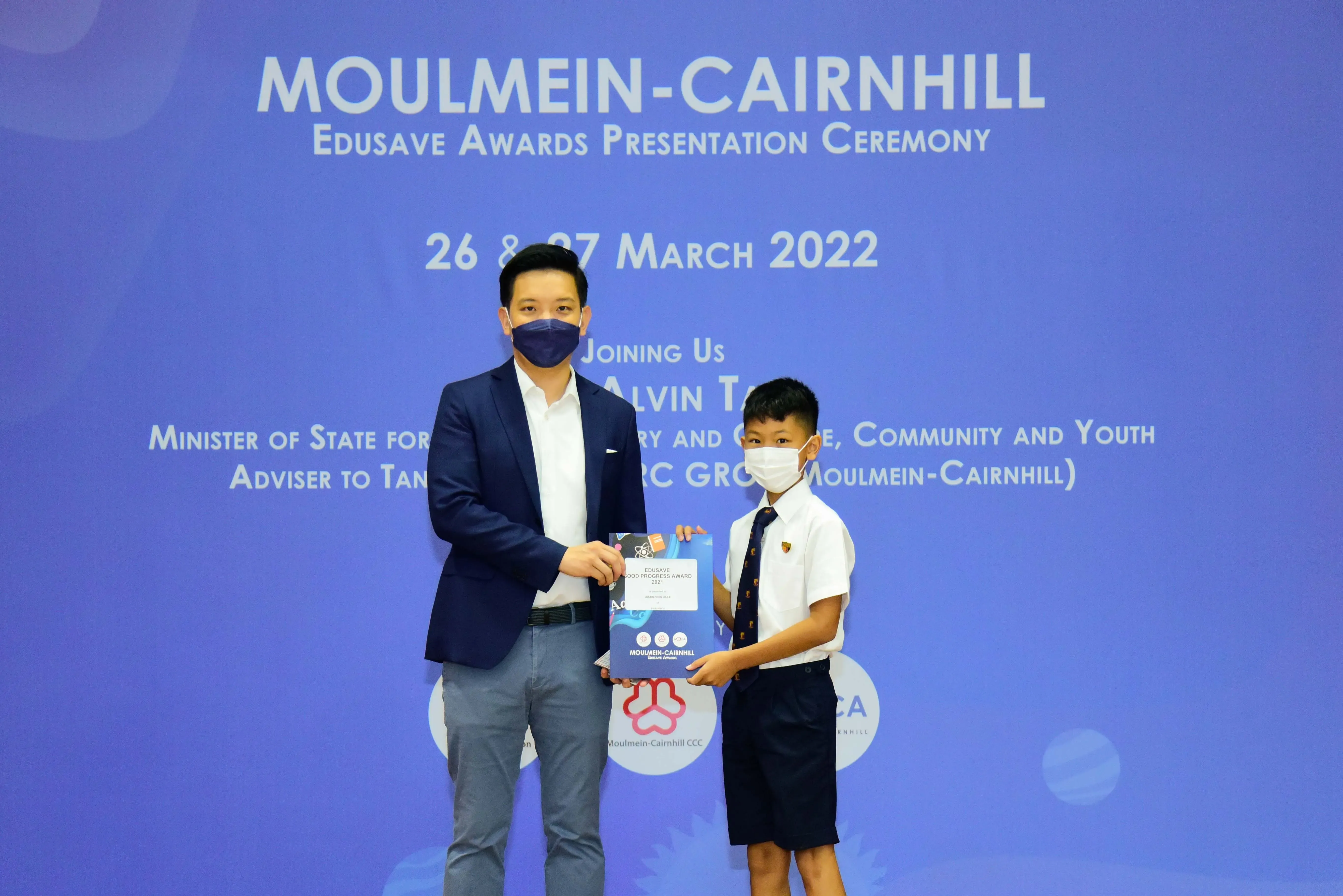 An adult in a suit and a young student in uniform hold an award at the Moulmein-Cairnhill Edusave ceremony, March 2022.