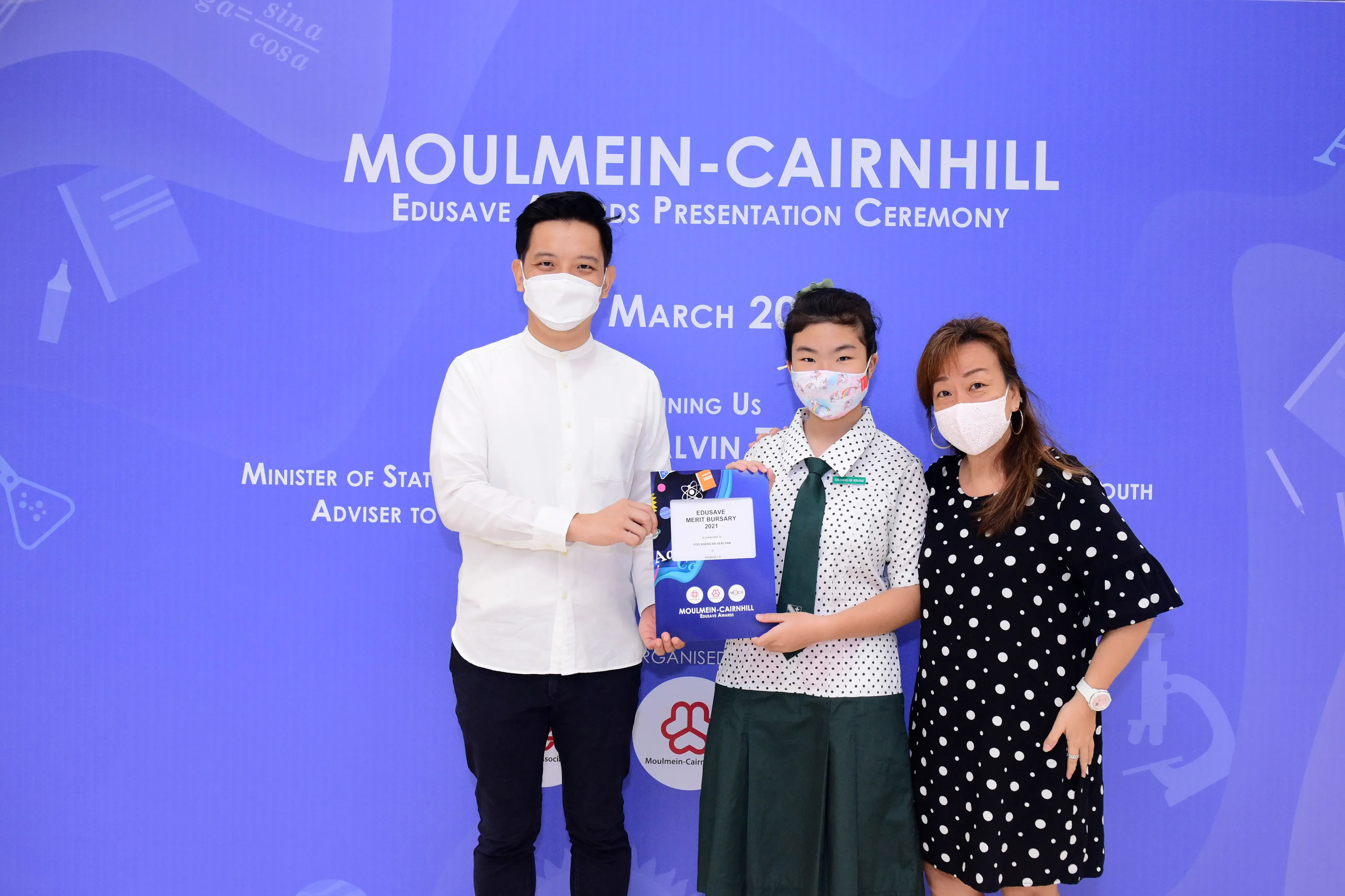 Three masked people pose at the Moulmein-Cairnhill Edusave Awards Ceremony, with a student holding an award certificate.