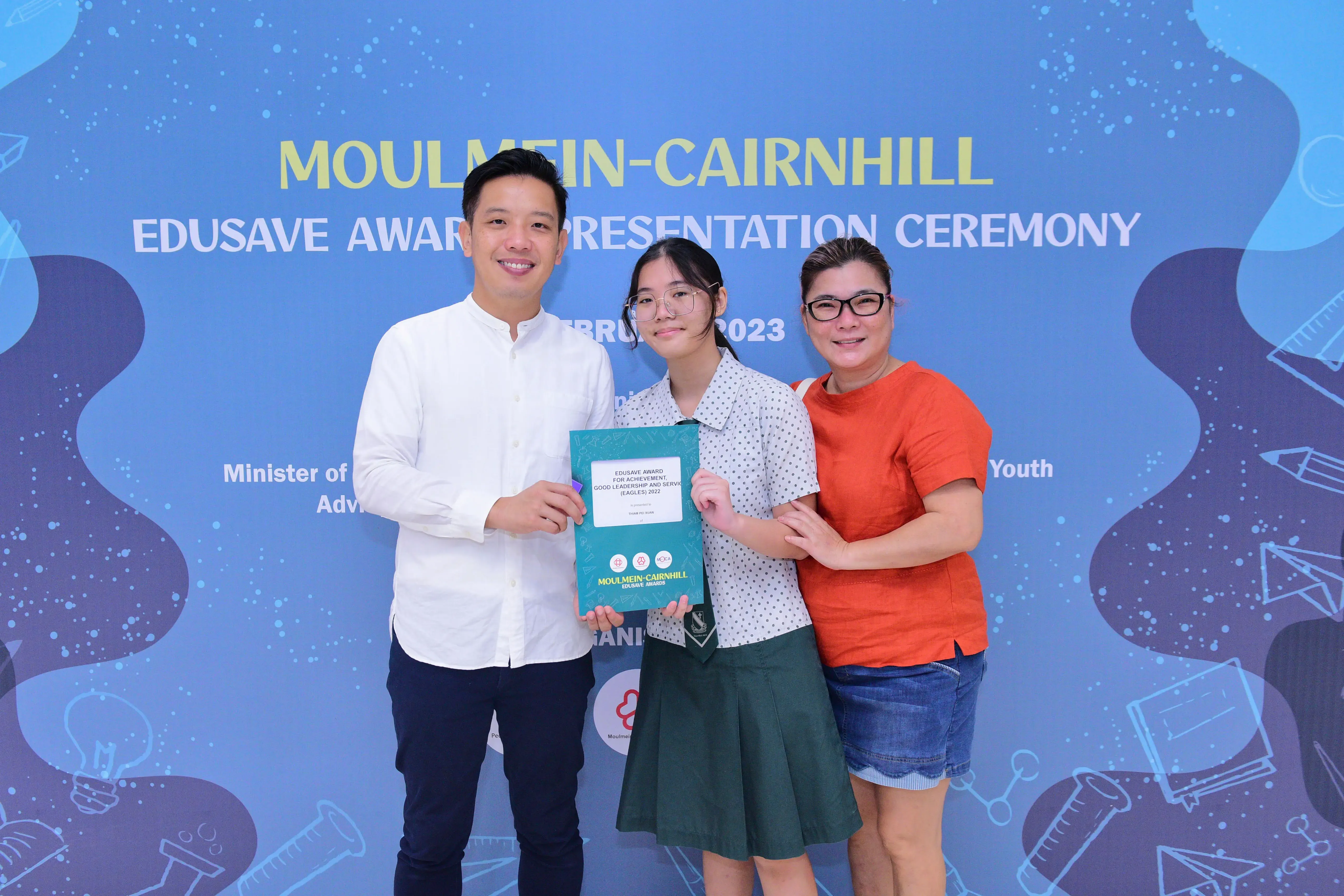 Three people smile and hold an award certificate at the Moulmein-Cairnhill Edusave Award Presentation Ceremony 2023.
