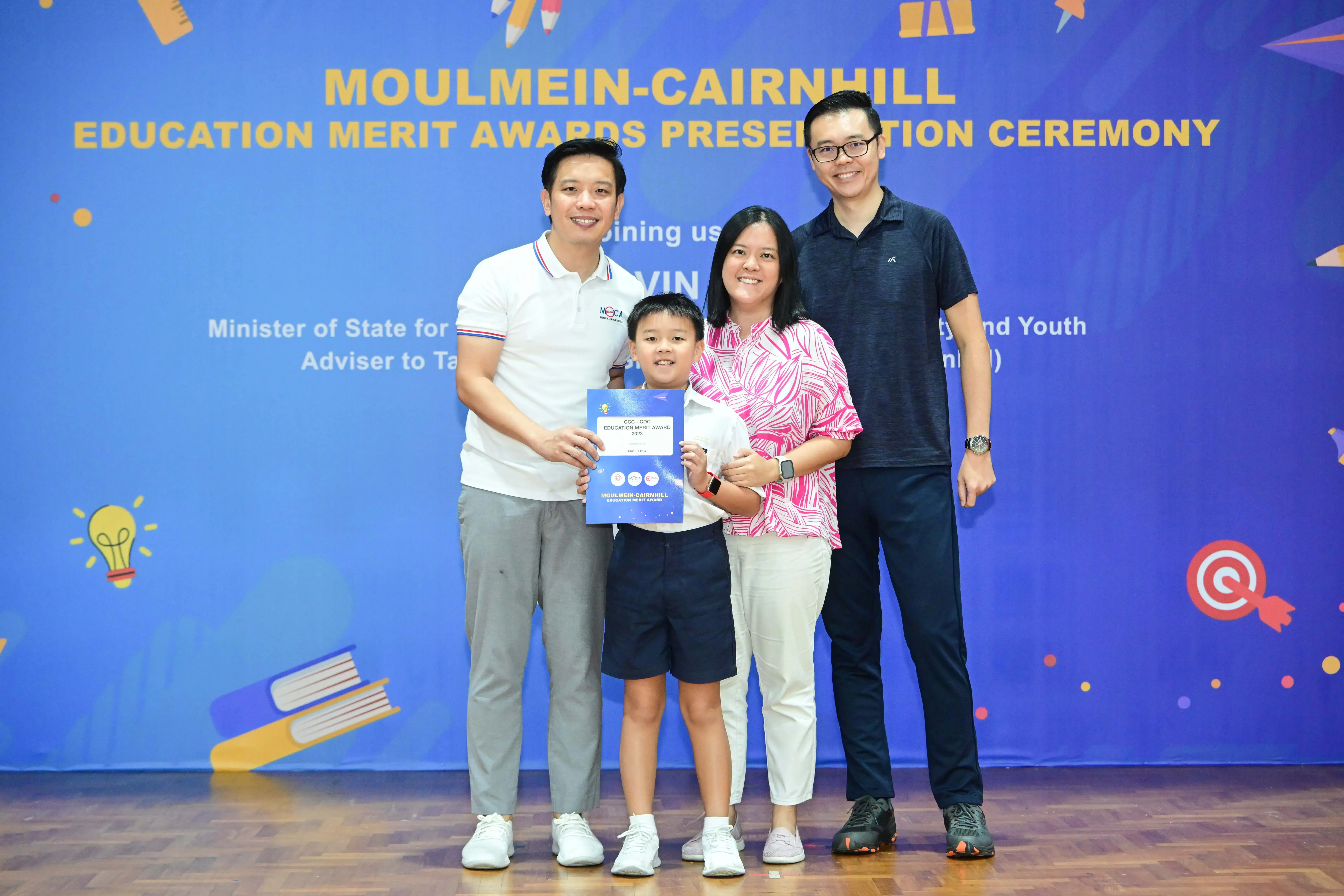 A boy holds an award certificate with three adults at the Moulmein-Cairnhill Education Merit Awards Presentation Ceremony.