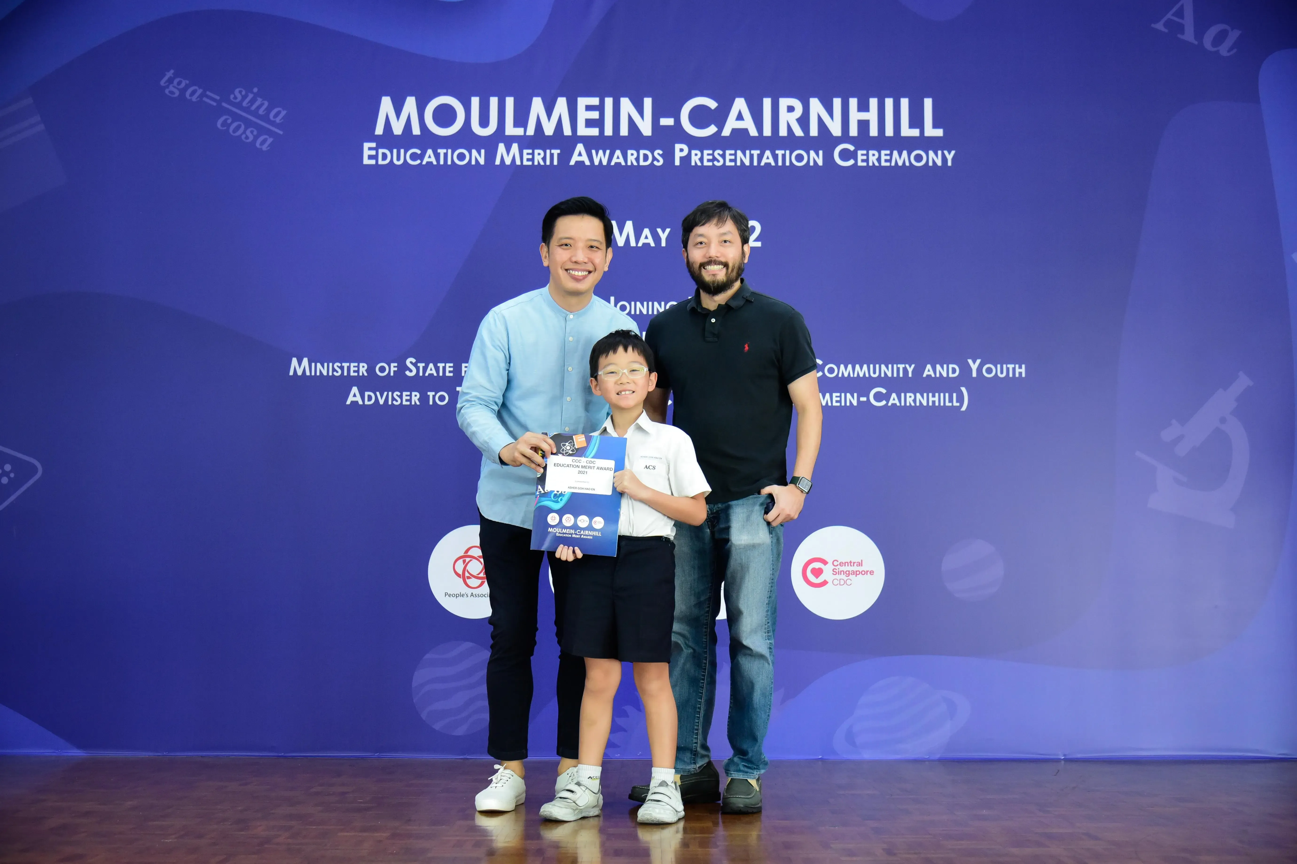 Two men and a young boy holding an award certificate pose at the Moulmein-Cairnhill Education Merit Awards ceremony.