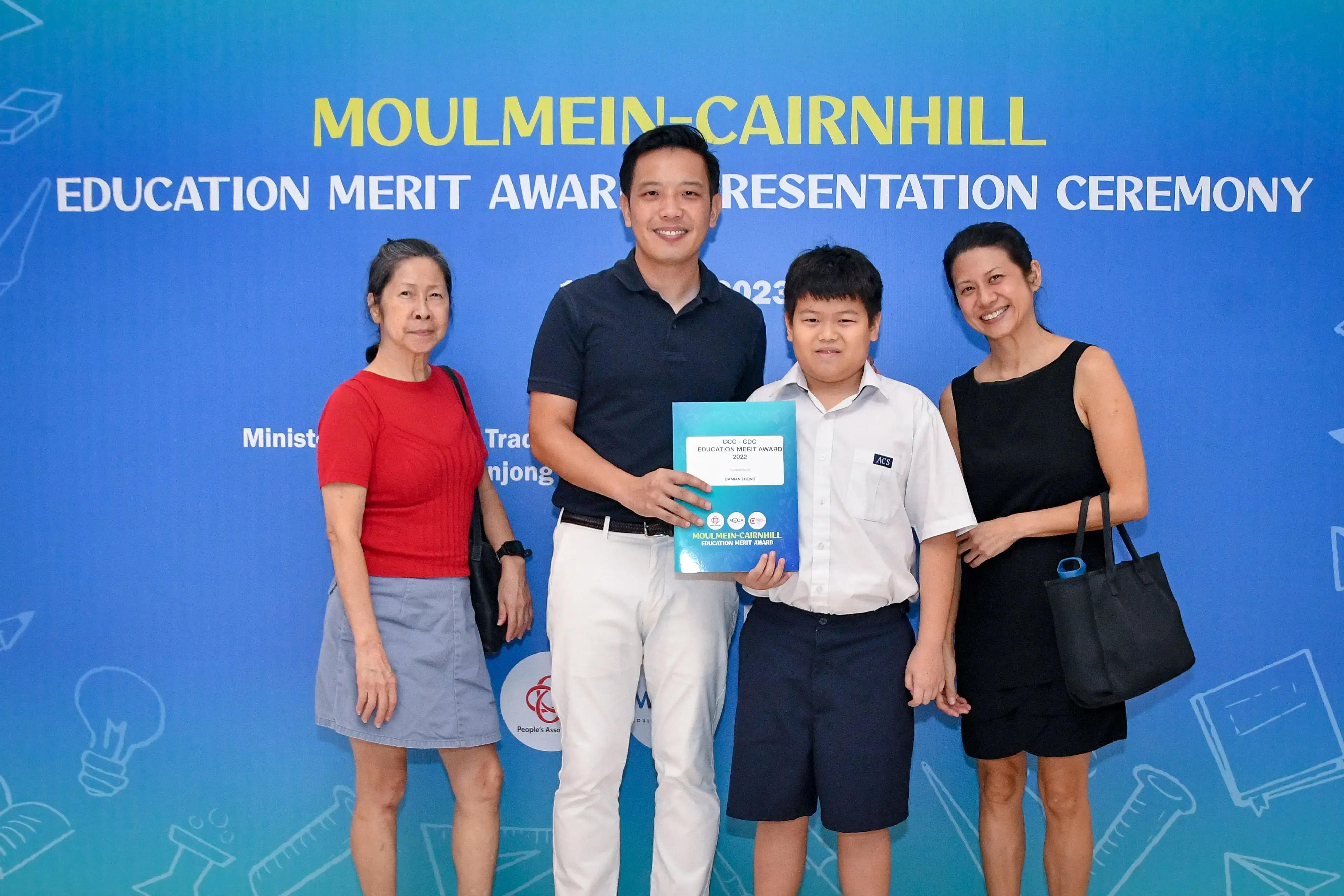 Four people pose before a Moulmein-Cairnhill Education Merit Award Presentation Ceremony banner, a boy holding an award.