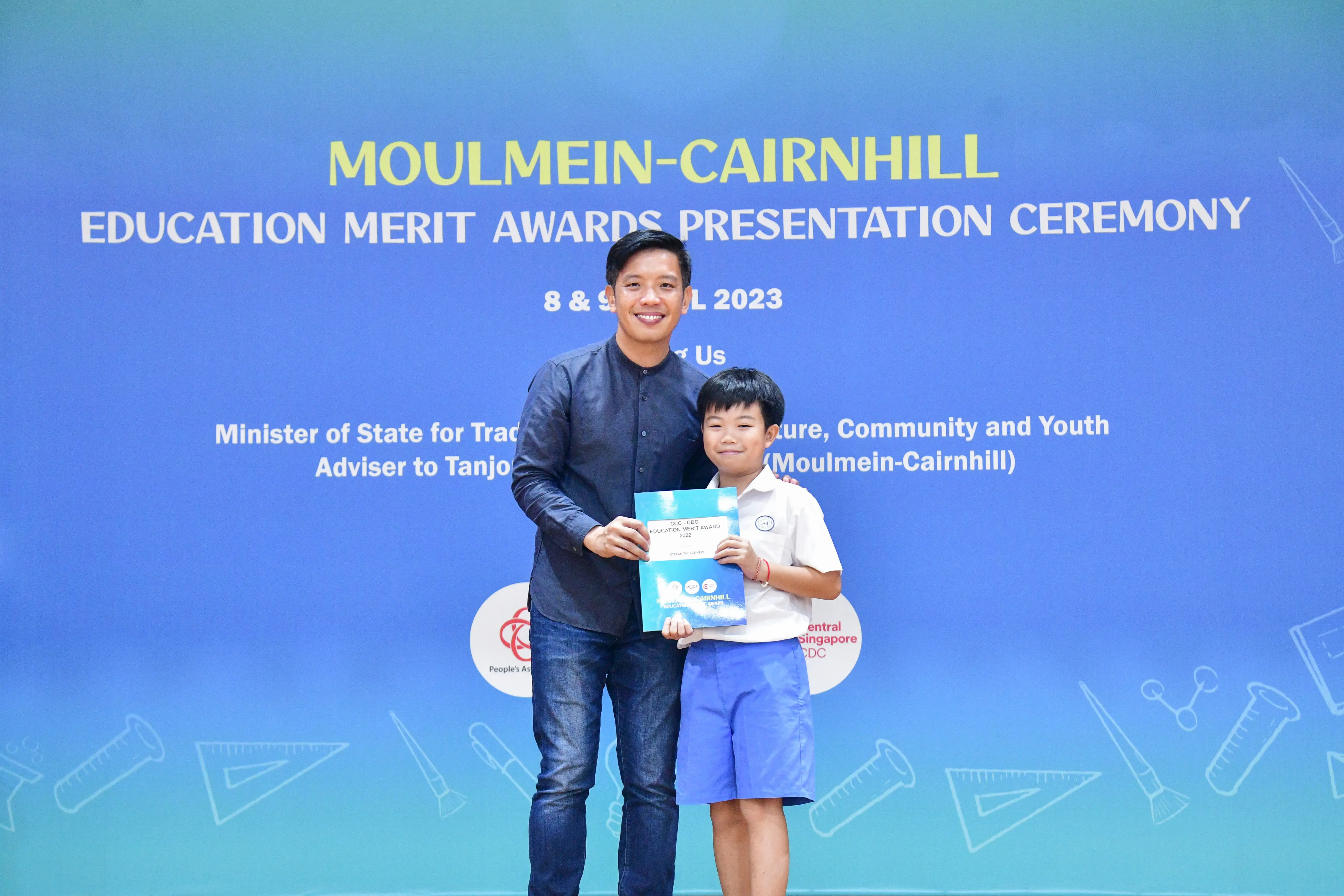 A man and a young boy hold a certificate in front of a Moulmein-Cairnhill Education Merit Awards banner.