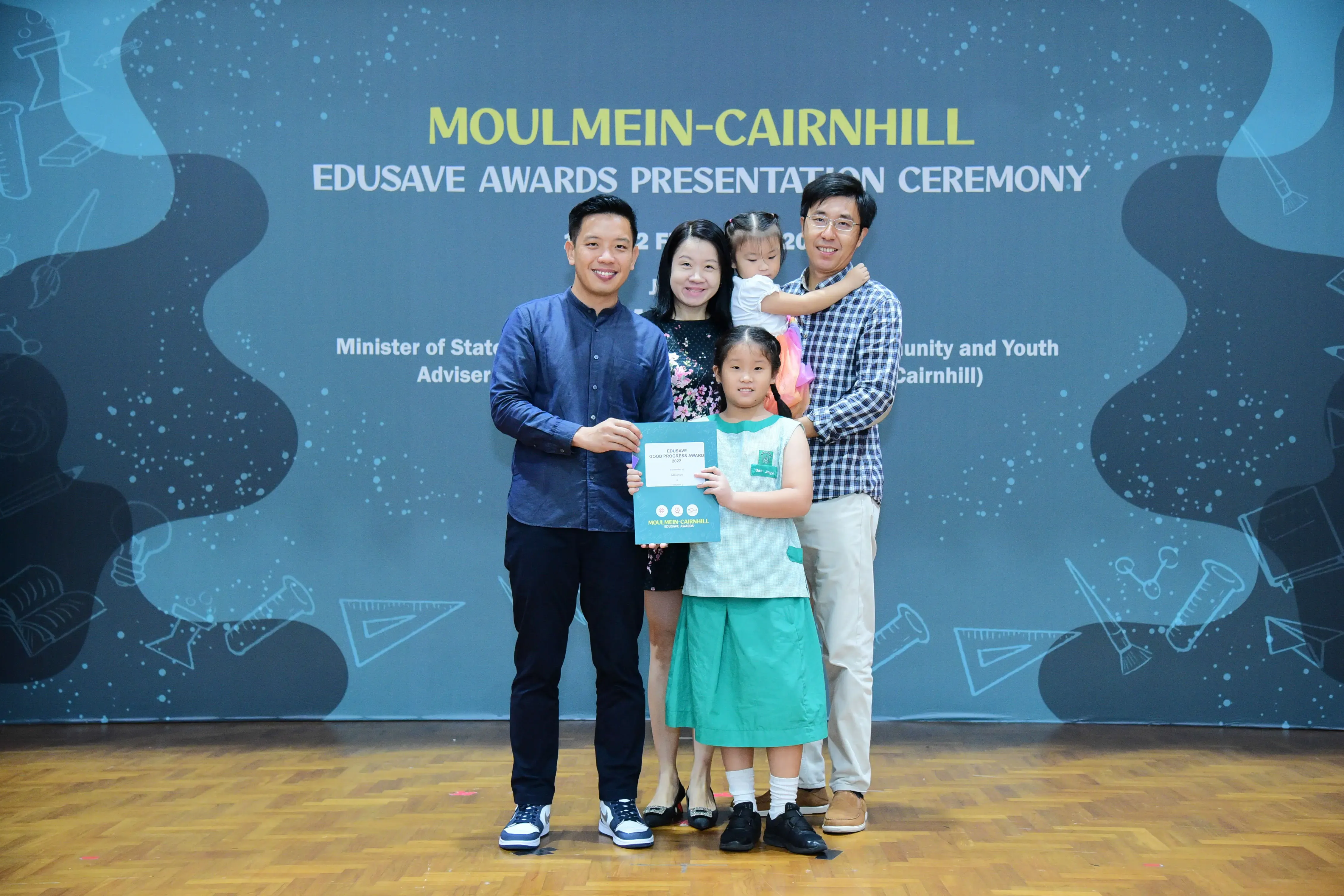 A girl holds a certificate with three adults and a toddler in front of a Moulmein-Cairnhill Edusave Awards banner.