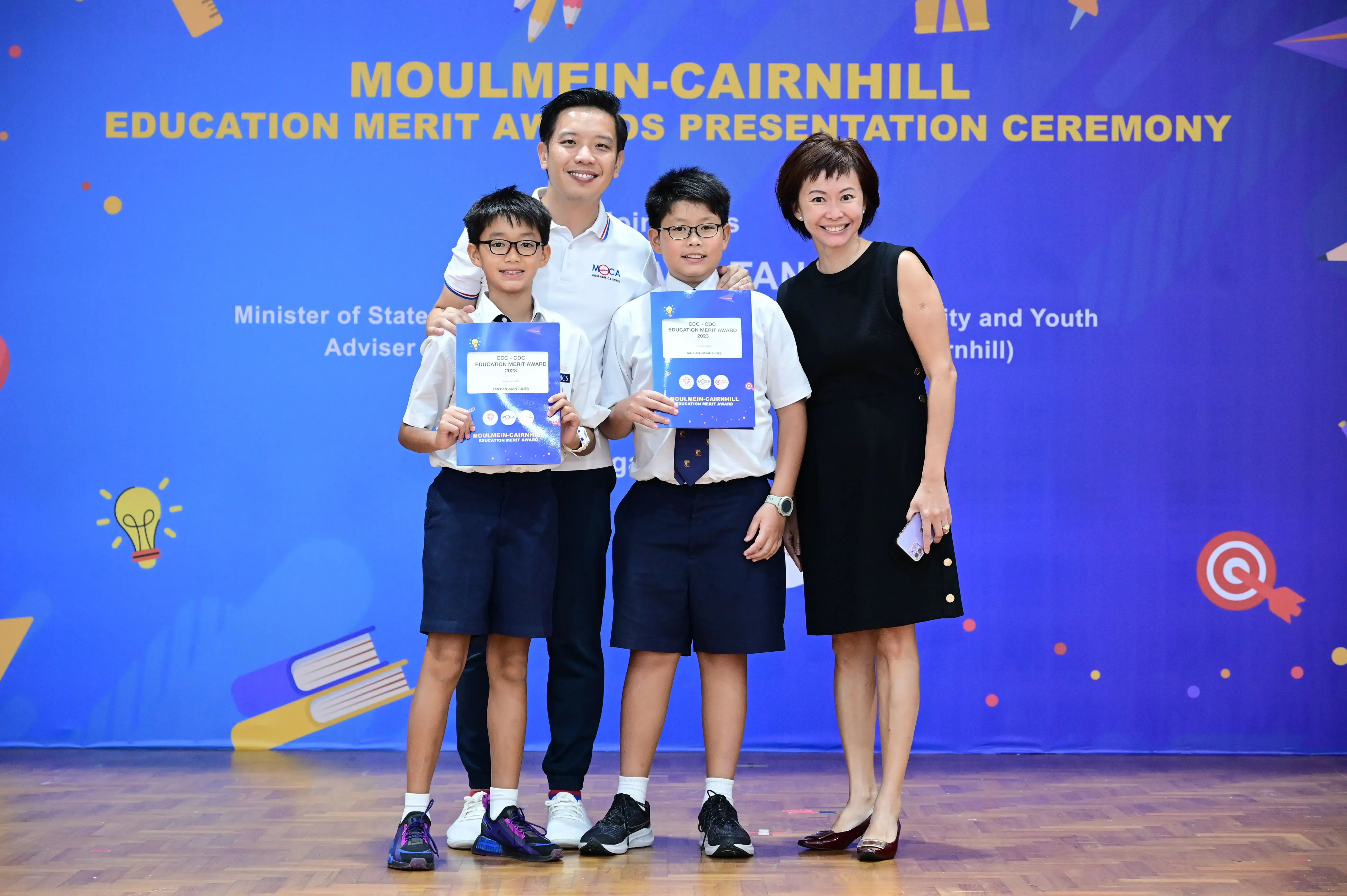 Two adults and two boys in school uniforms hold award certificates at the Moulmein-Cairnhill Education Merit Awards ceremony.