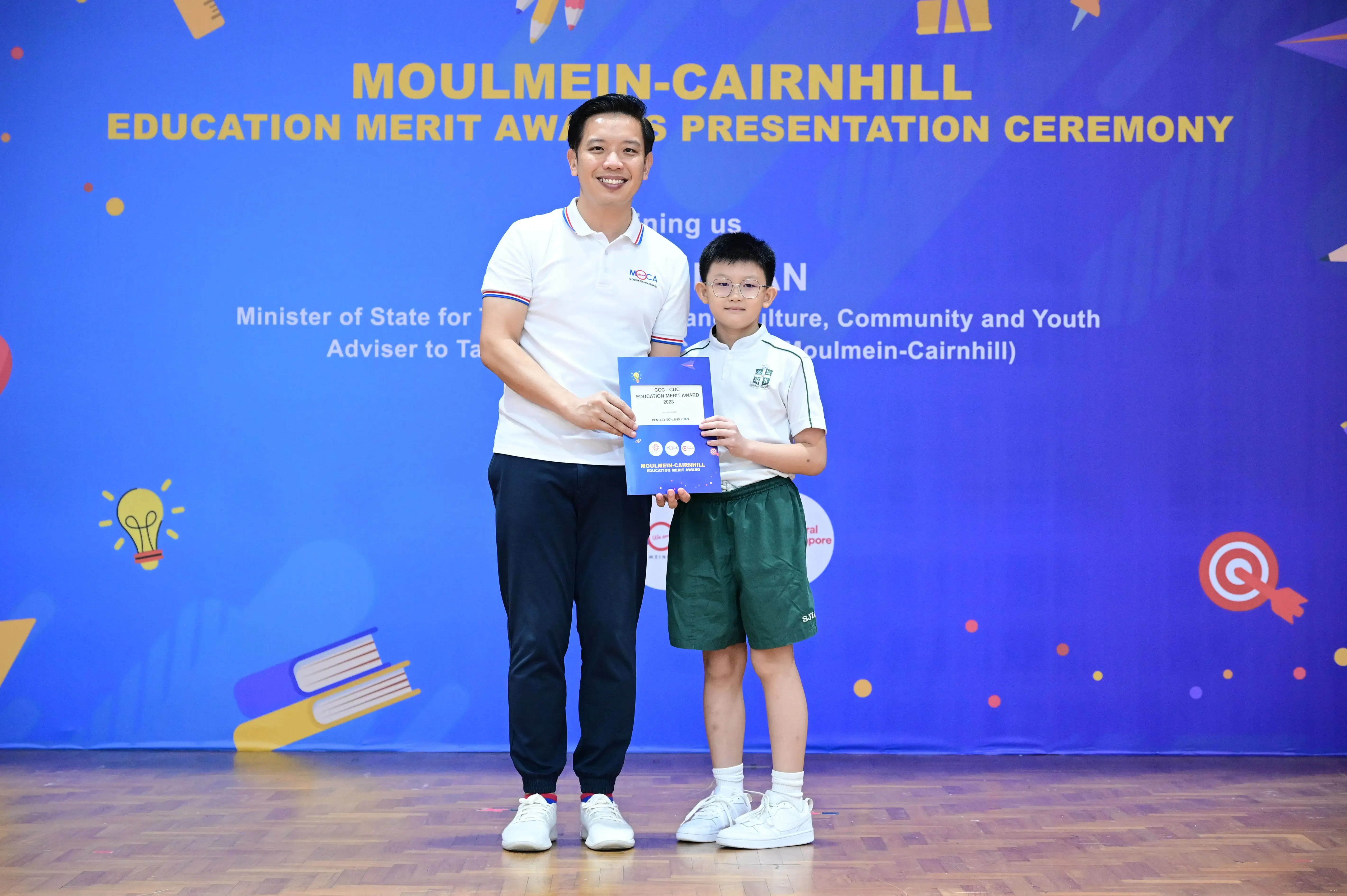 An adult man and a young boy hold a merit award certificate at the Moulmein-Cairnhill Education Merit Awards ceremony.