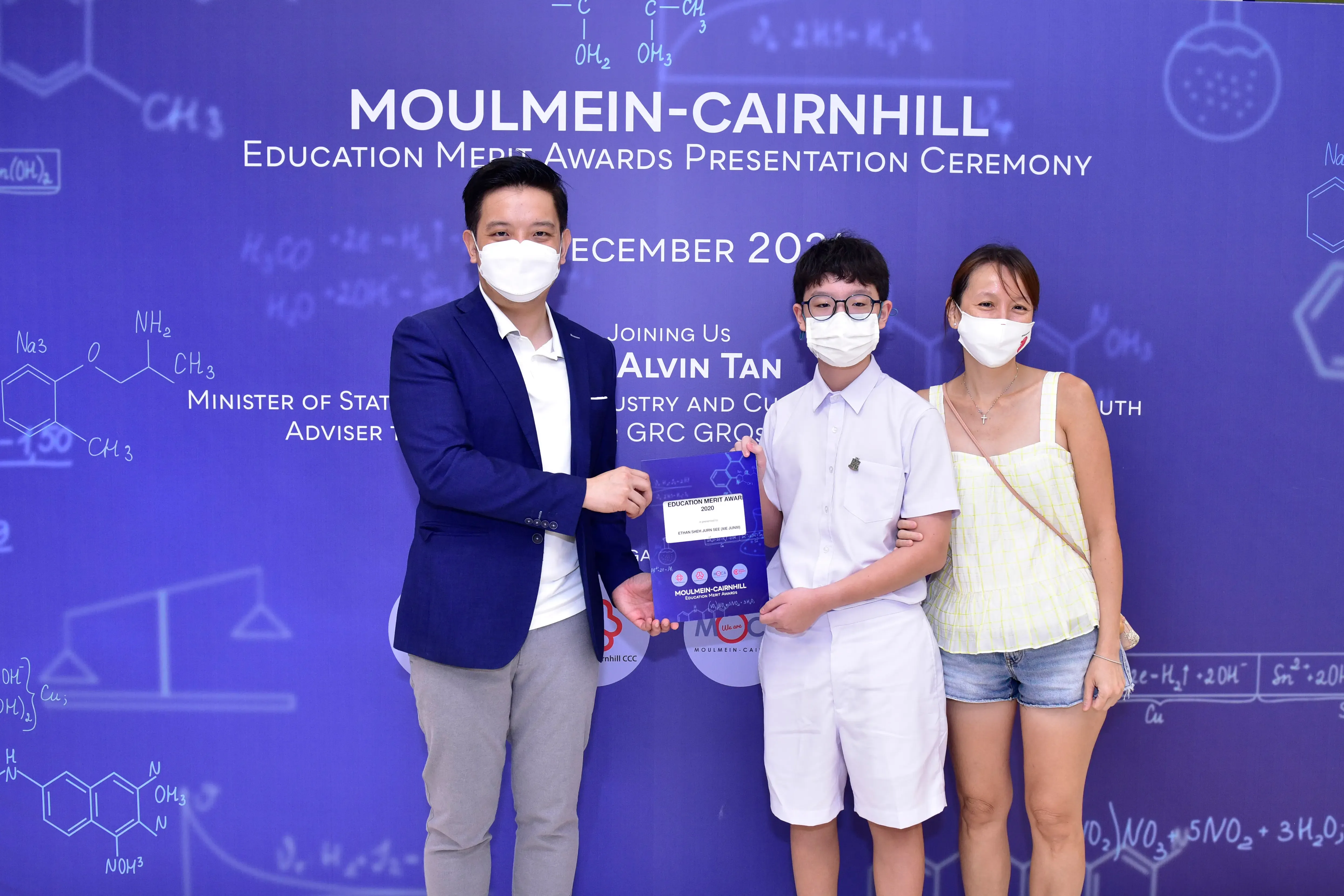 Three masked people pose at Moulmein-Cairnhill Education Merit Awards ceremony, with a boy holding an award certificate.