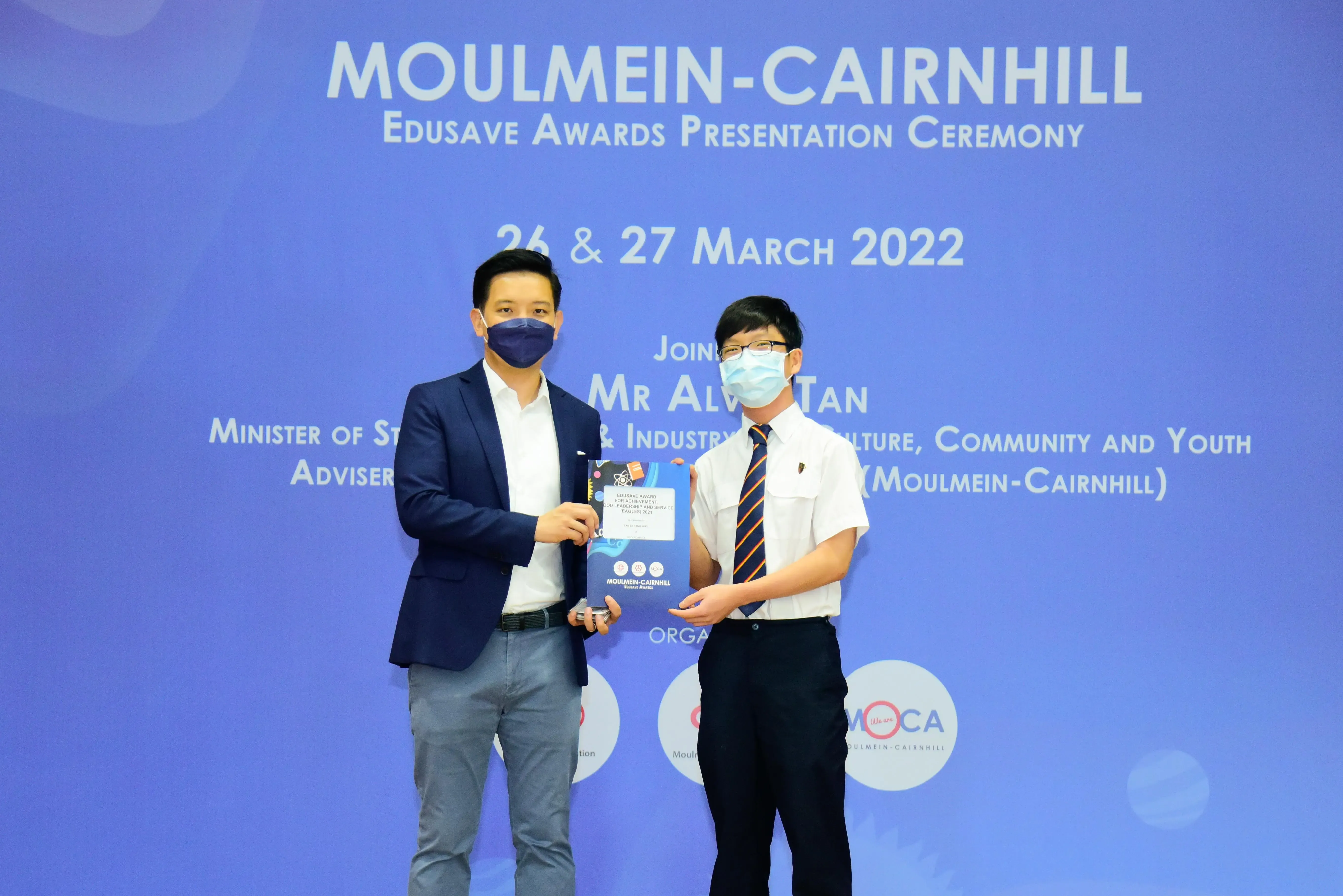 Two masked men hold an award folder at the Moulmein-Cairnhill Edusave Awards Presentation Ceremony, March 2022.