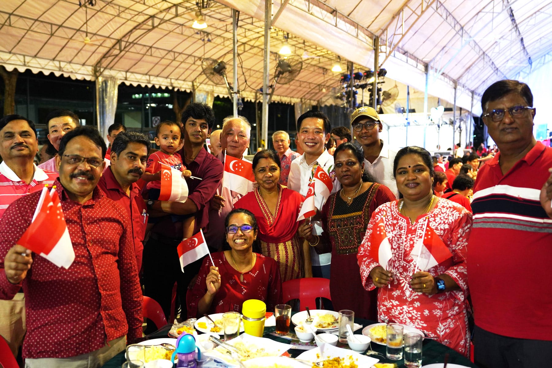 A diverse group of smiling people in red clothing wave Singapore flags at an outdoor tent event with food on tables.