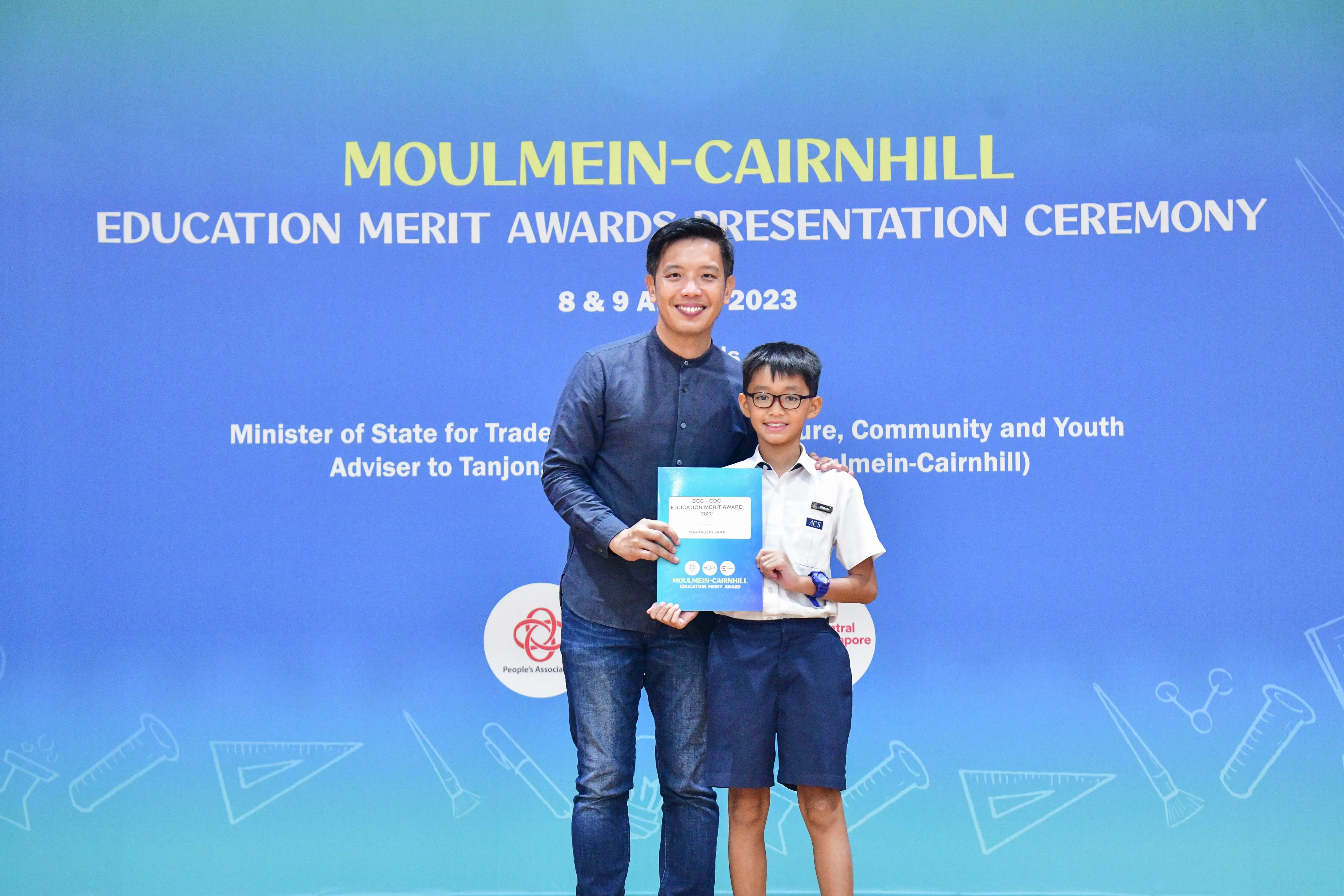Adult man and young boy holding an award certificate at the Moulmein-Cairnhill Education Merit Awards Ceremony 2023.