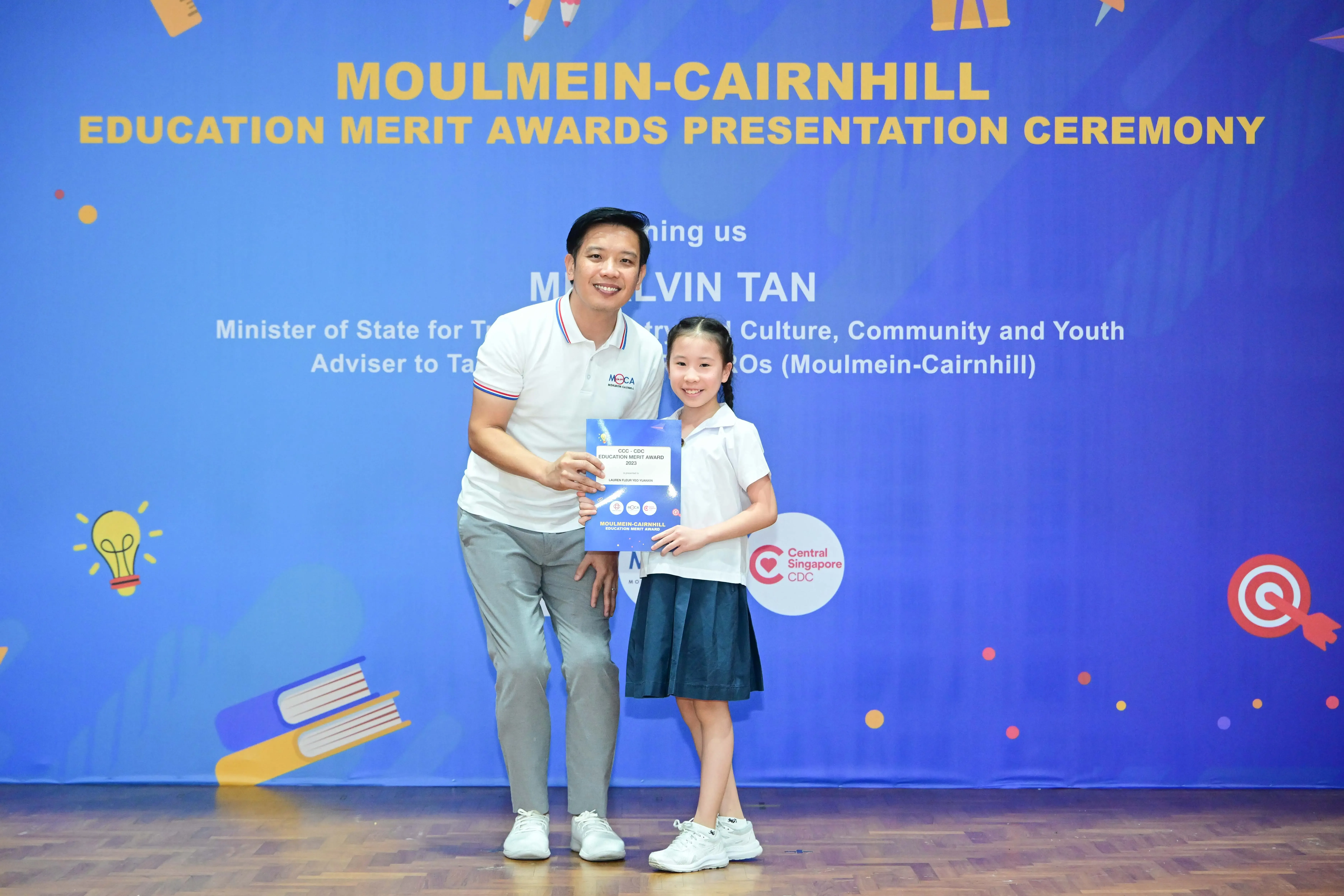 A man and a young girl hold an award certificate in front of a Moulmein-Cairnhill Education Merit Awards banner.
