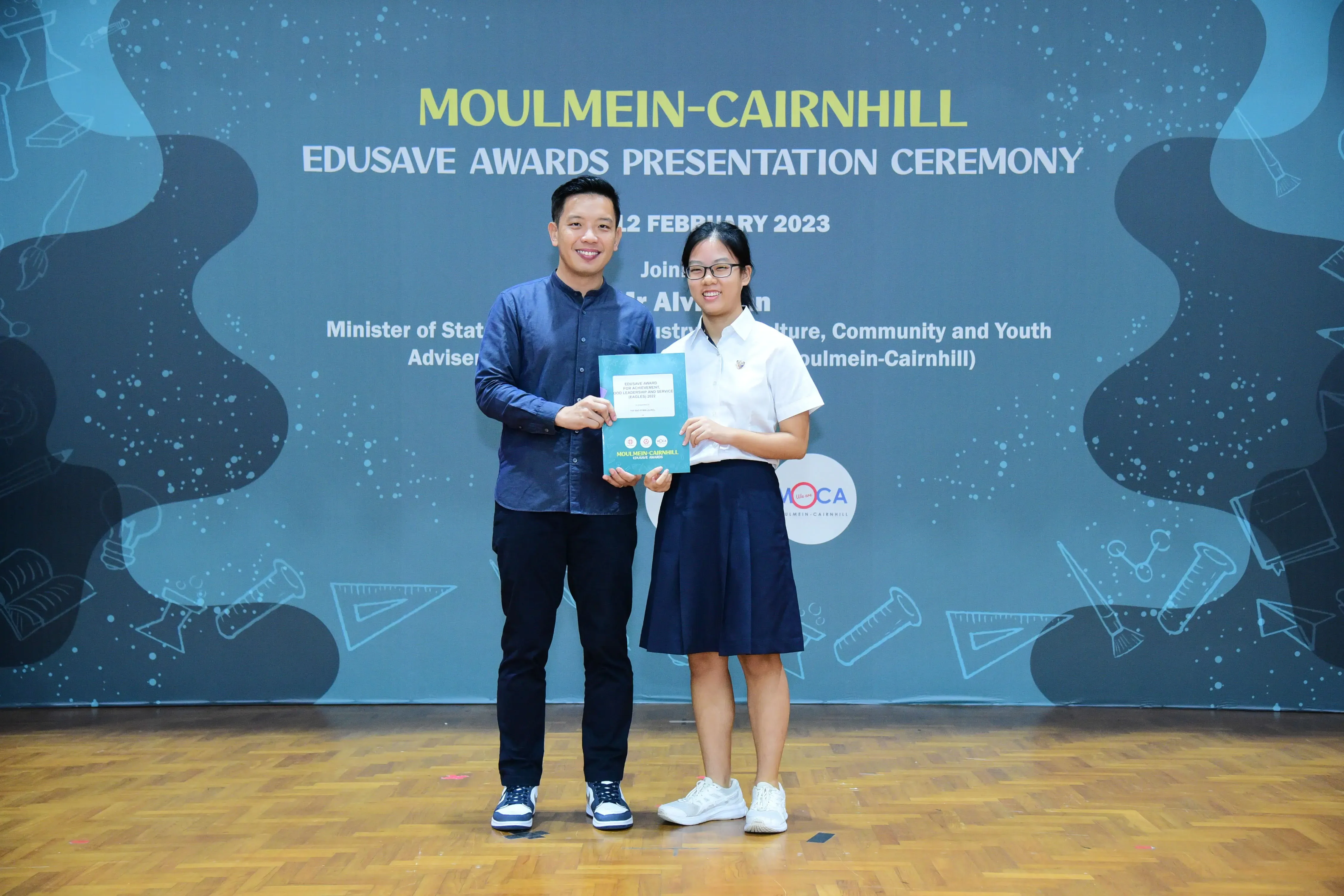 Two people hold a certificate together on stage at the Moulmein-Cairnhill Edusave Awards Presentation Ceremony, February 2023.