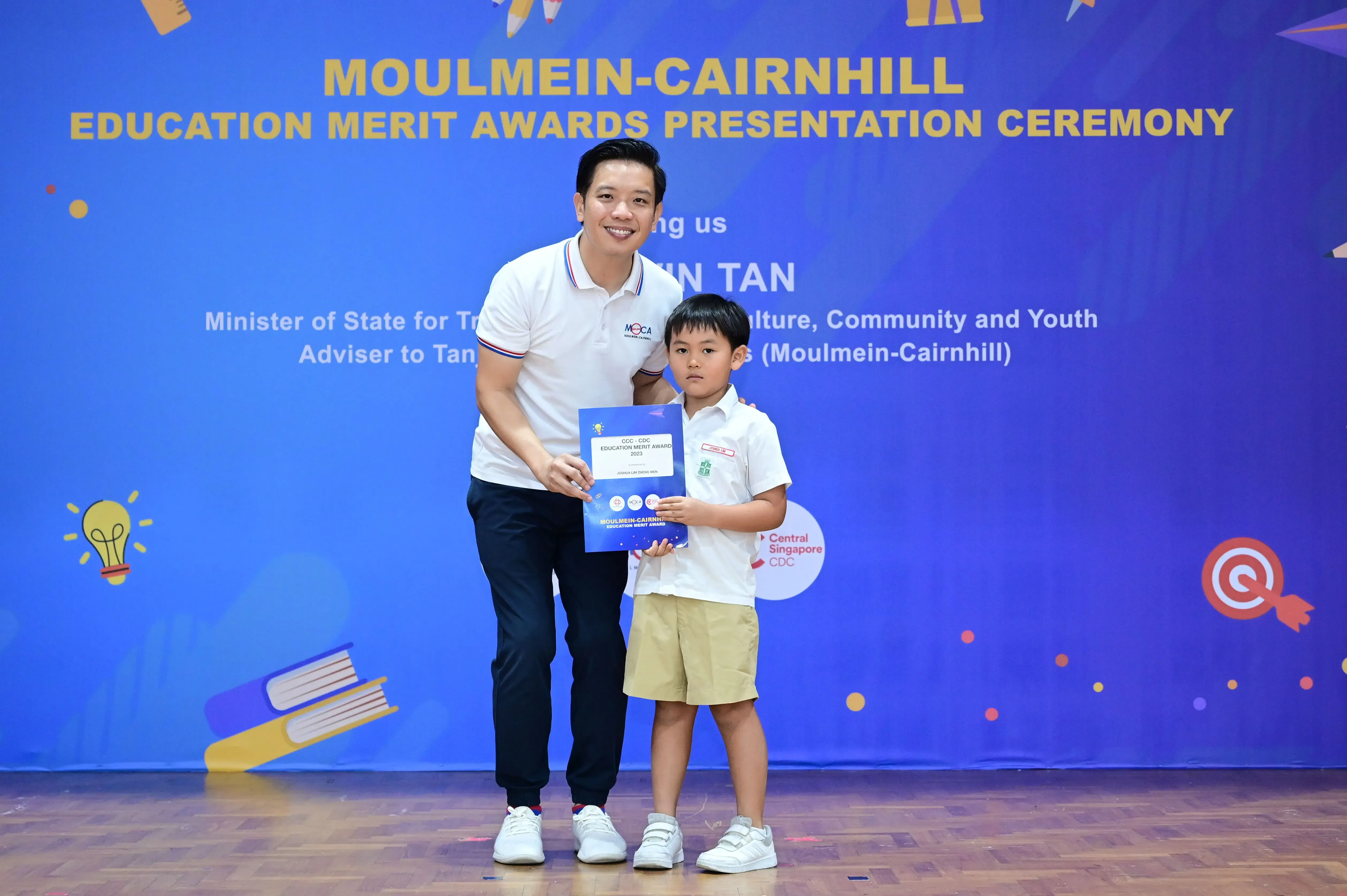 An adult and a young boy hold a certificate in front of a Moulmein-Cairnhill Education Merit Awards banner.