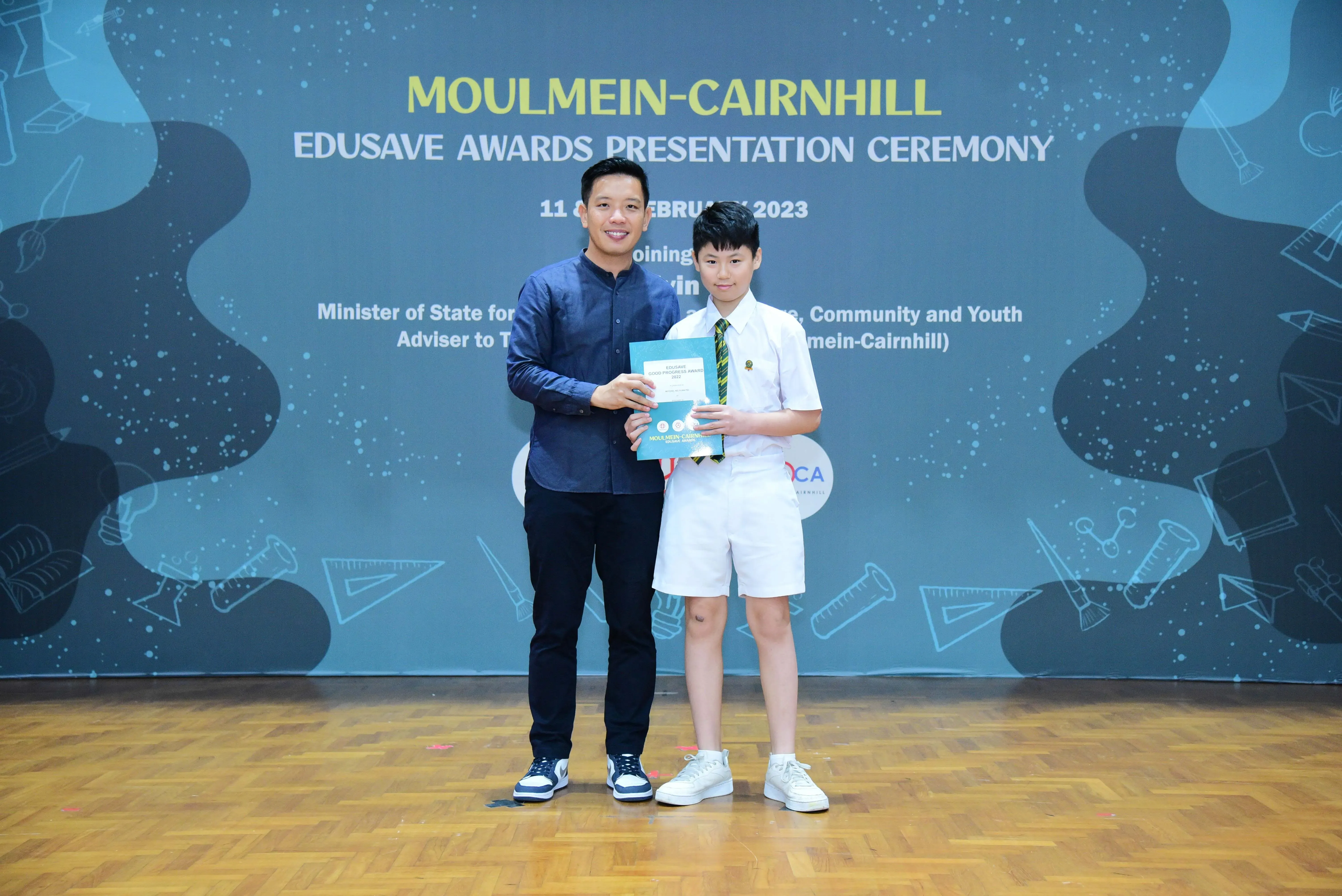 An adult and a young boy in school uniform hold a certificate at the Moulmein-Cairnhill Edusave Awards Ceremony 2023.
