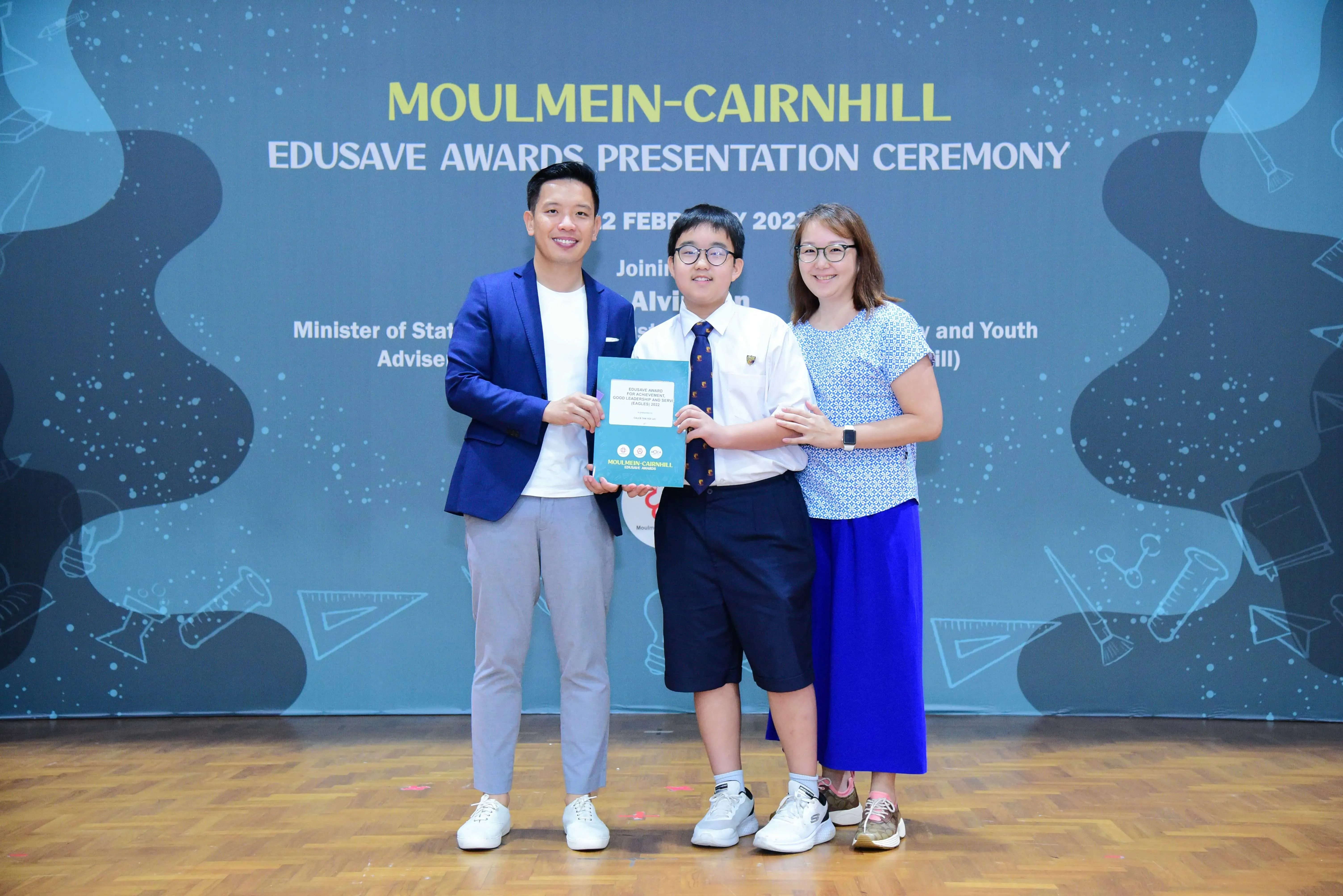 Three people pose holding a certificate at the Moulmein-Cairnhill Edusave Awards Presentation Ceremony backdrop.