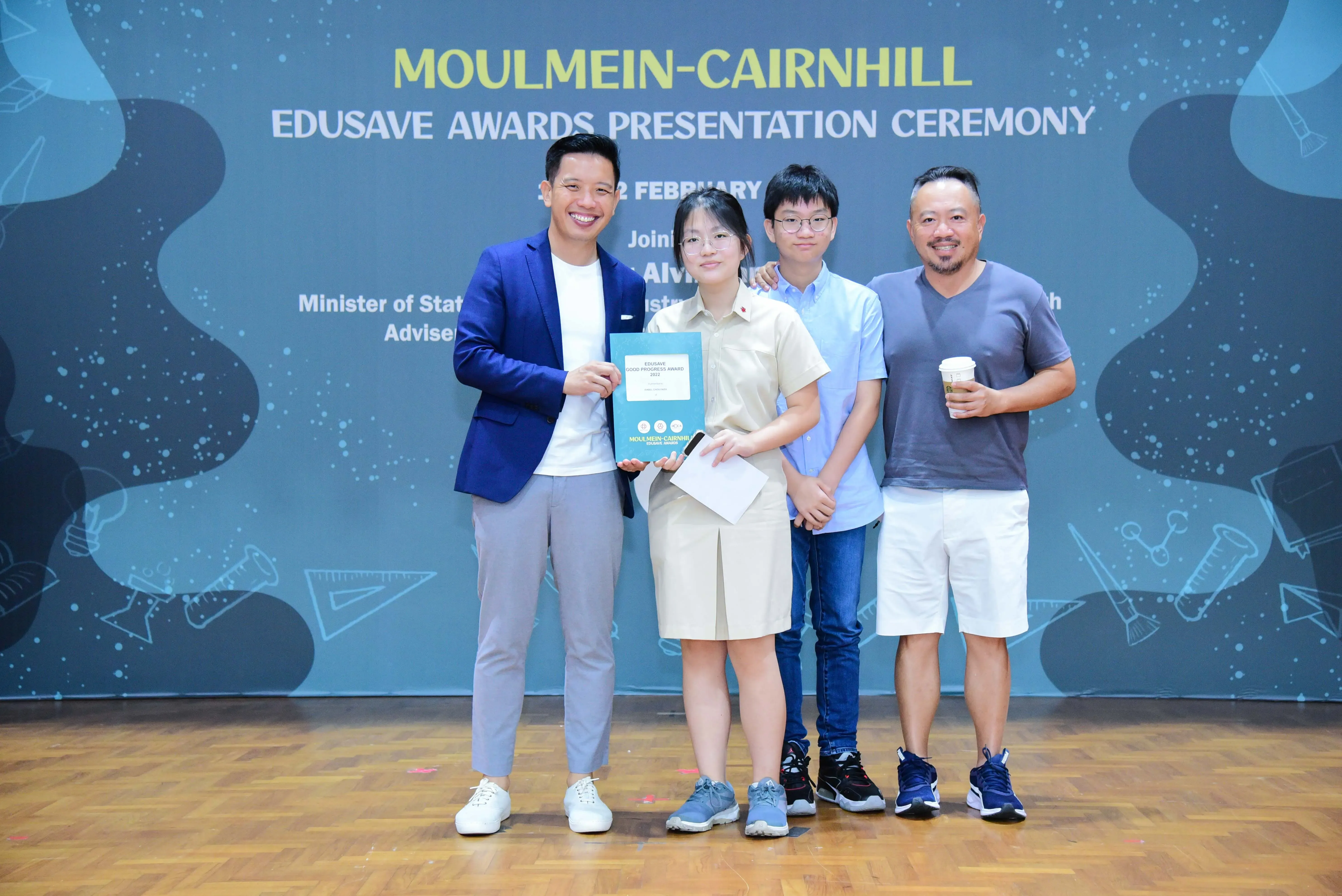Four people pose at the Moulmein-Cairnhill Edusave Awards Presentation Ceremony, with a girl holding an award folder.