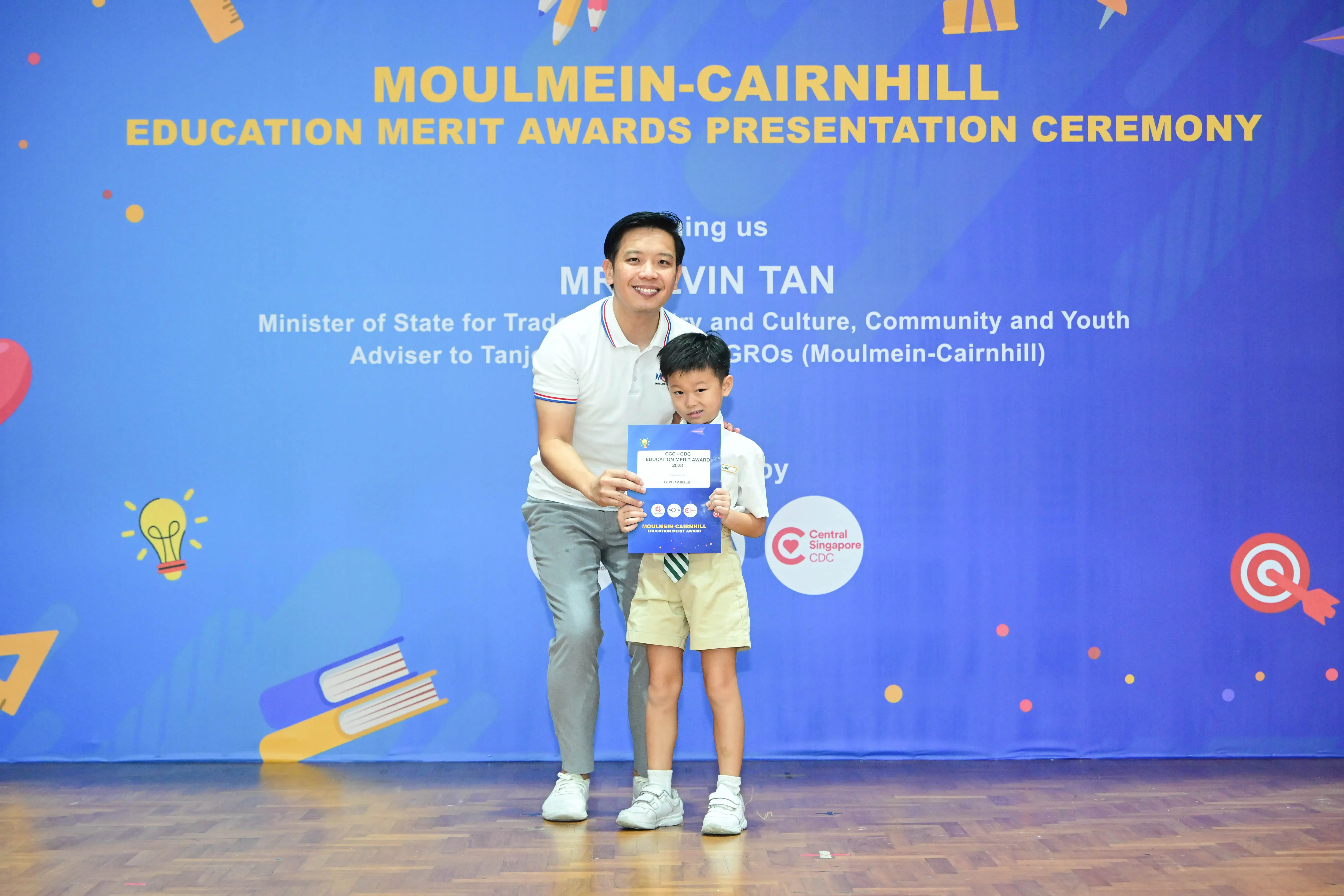 A man and young boy hold a certificate in front of a Moulmein-Cairnhill Education Merit Awards Presentation Ceremony banner.