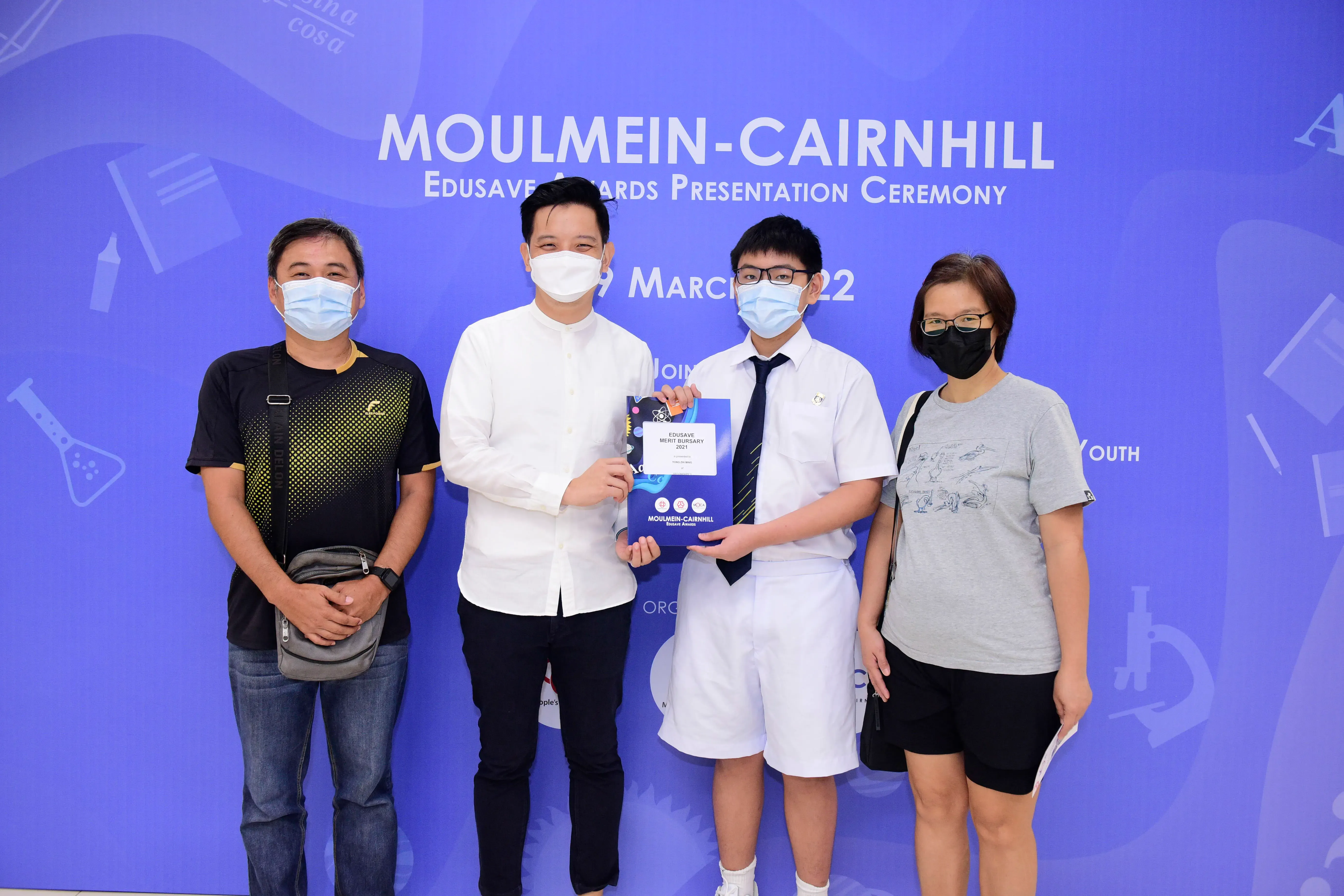 Four masked people pose at the Moulmein-Cairnhill Edusave Awards Presentation Ceremony, with a student holding a certificate.