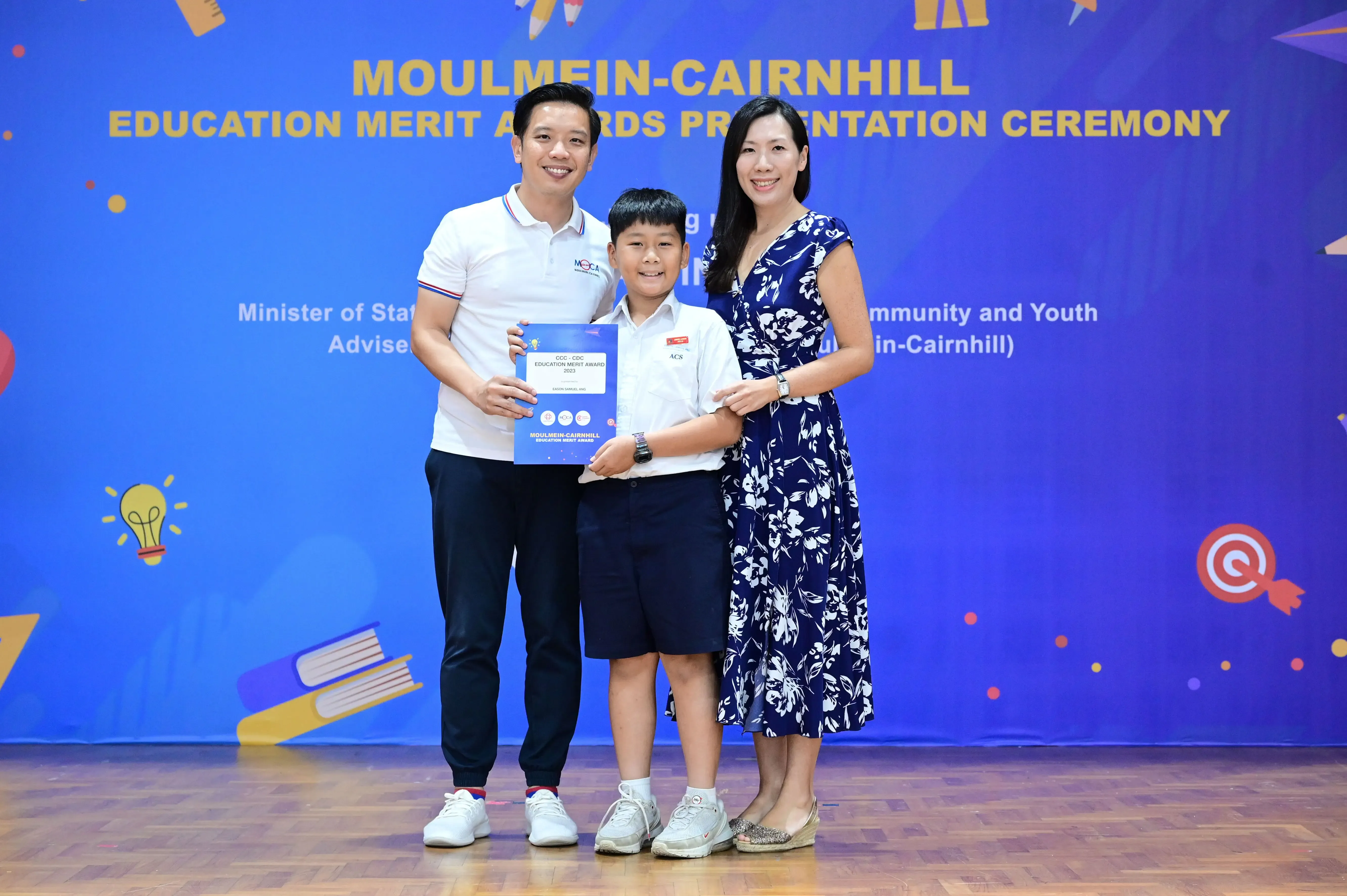 A boy holding an award certificate stands between two adults at the Moulmein-Cairnhill Education Merit Awards ceremony.