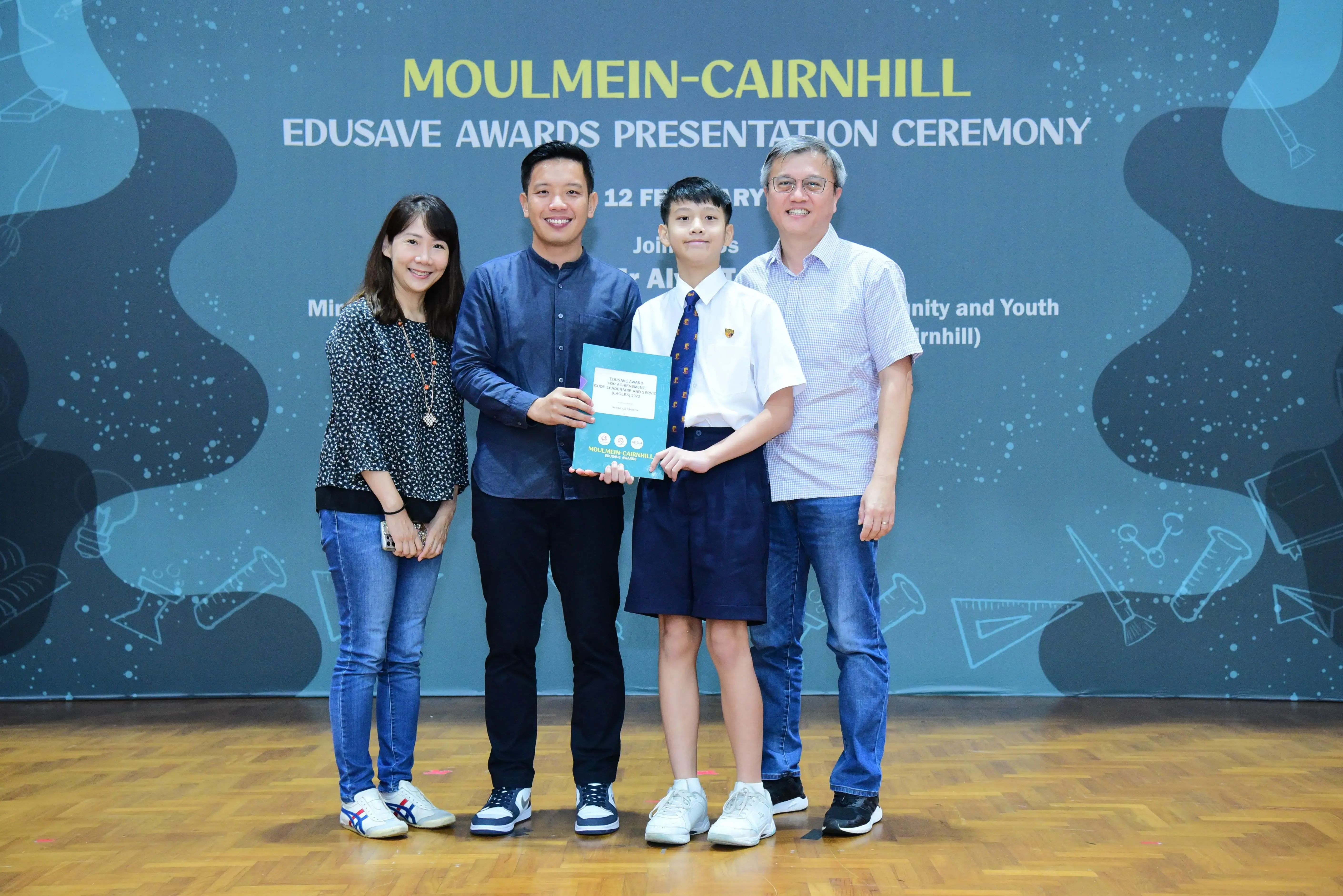 Four people pose at the Moulmein-Cairnhill Edusave Awards Presentation Ceremony, with a student holding a certificate.
