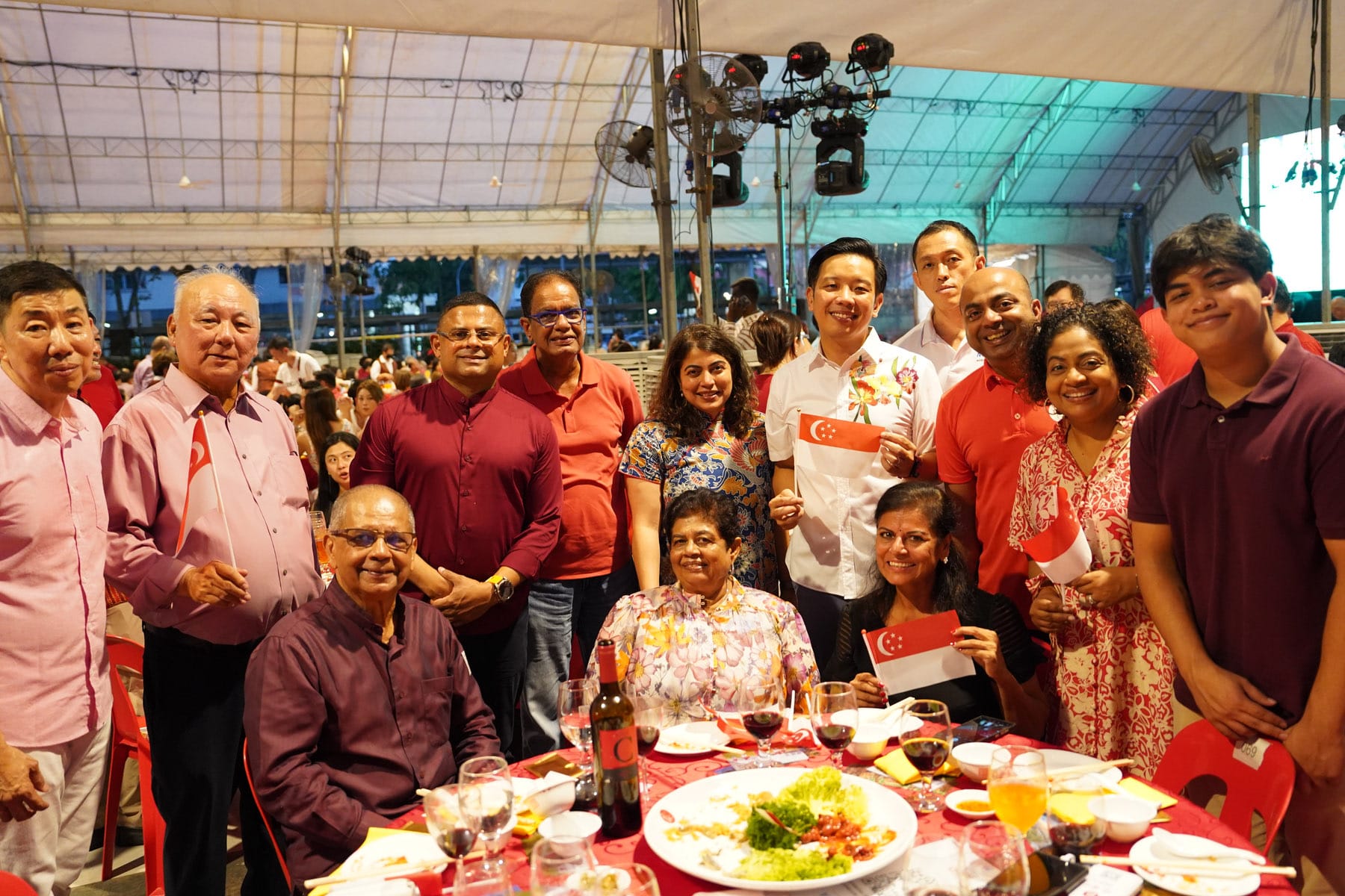 A group of about twelve people posing together at a festive outdoor dinner, some holding small Singapore flags.
