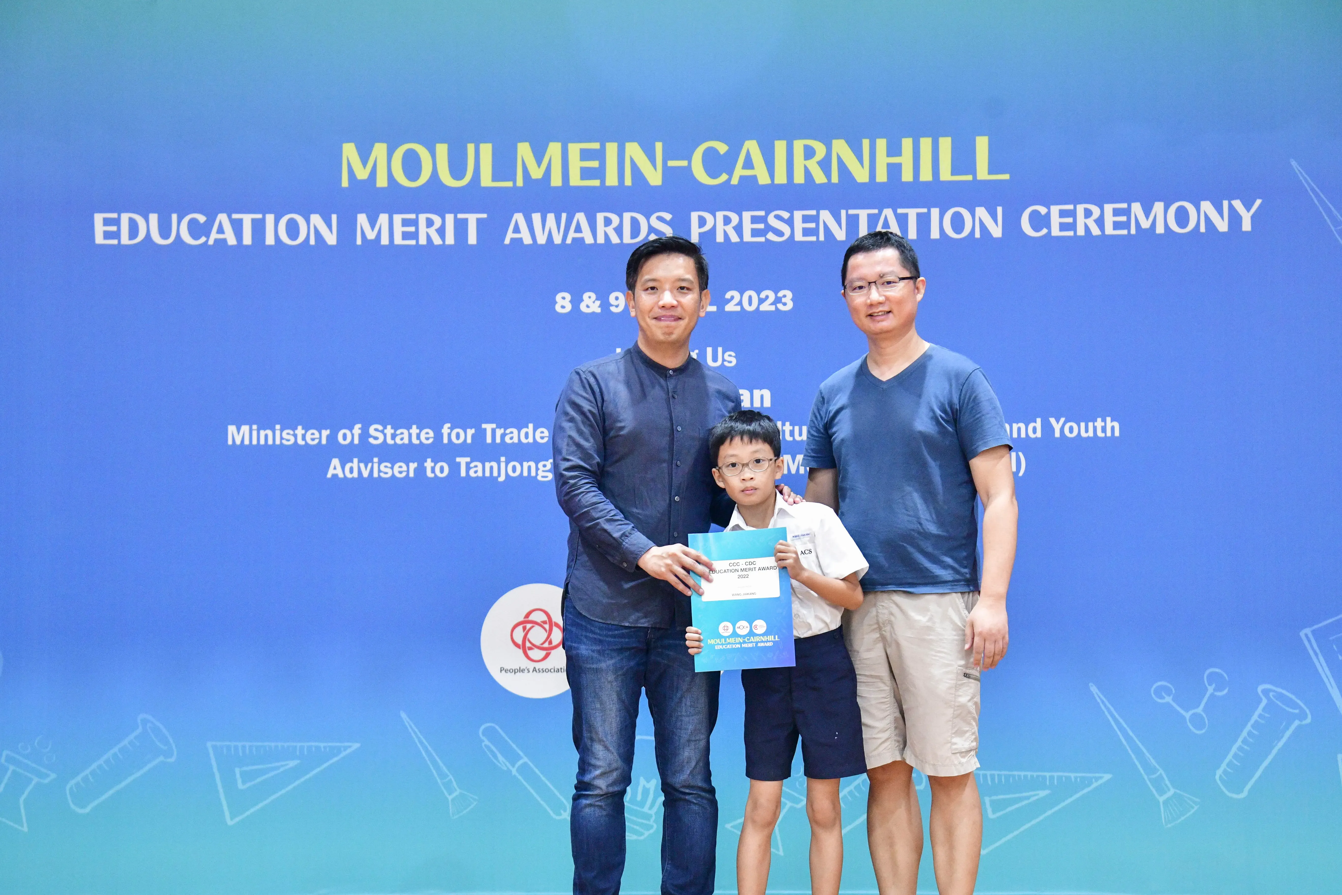 Two men and a boy holding an award certificate pose before a Moulmein-Cairnhill Education Merit Awards Ceremony banner.