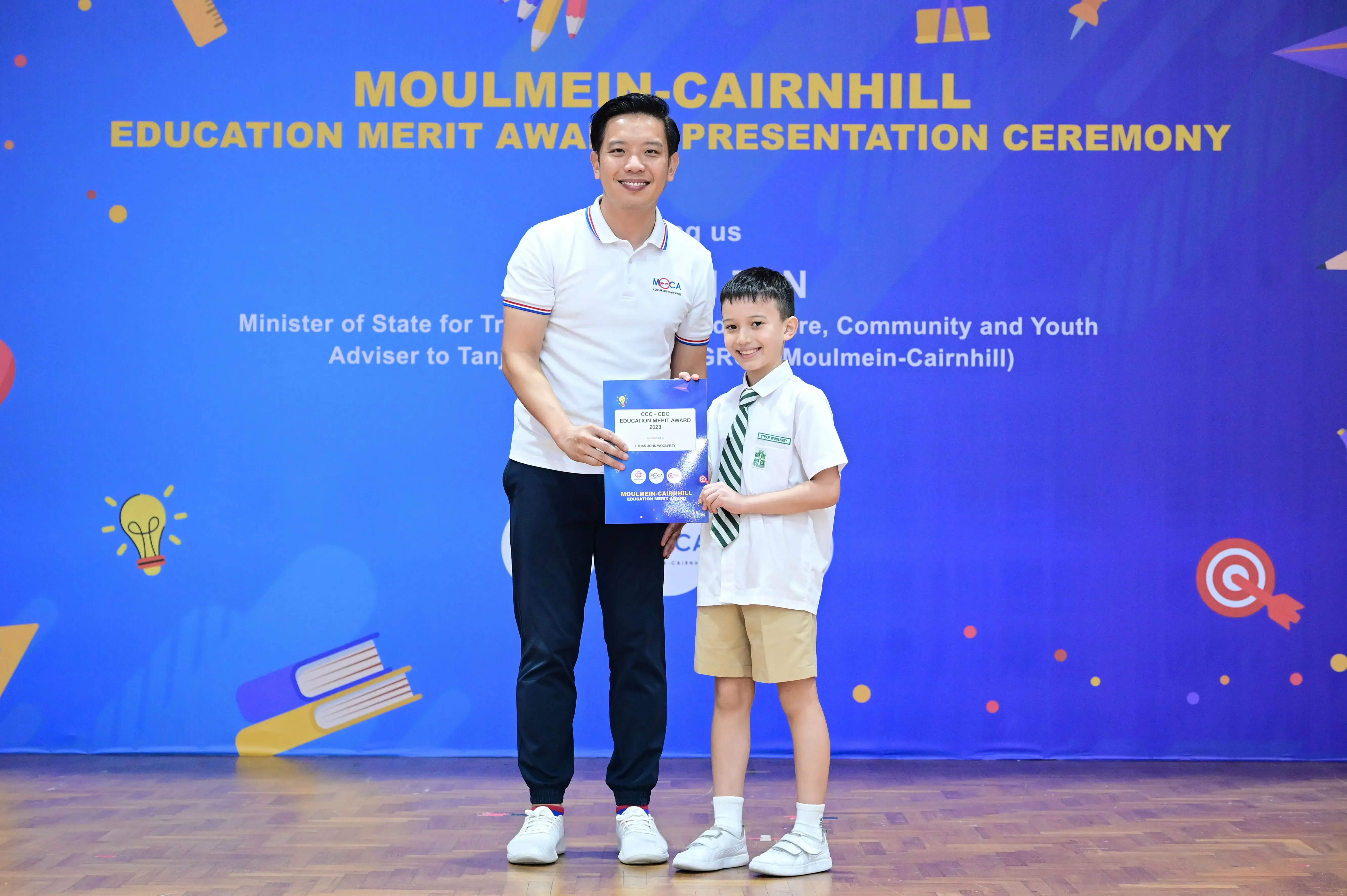 A man and a young boy in school uniform hold a certificate at the Moulmein-Cairnhill Education Merit Award Ceremony.