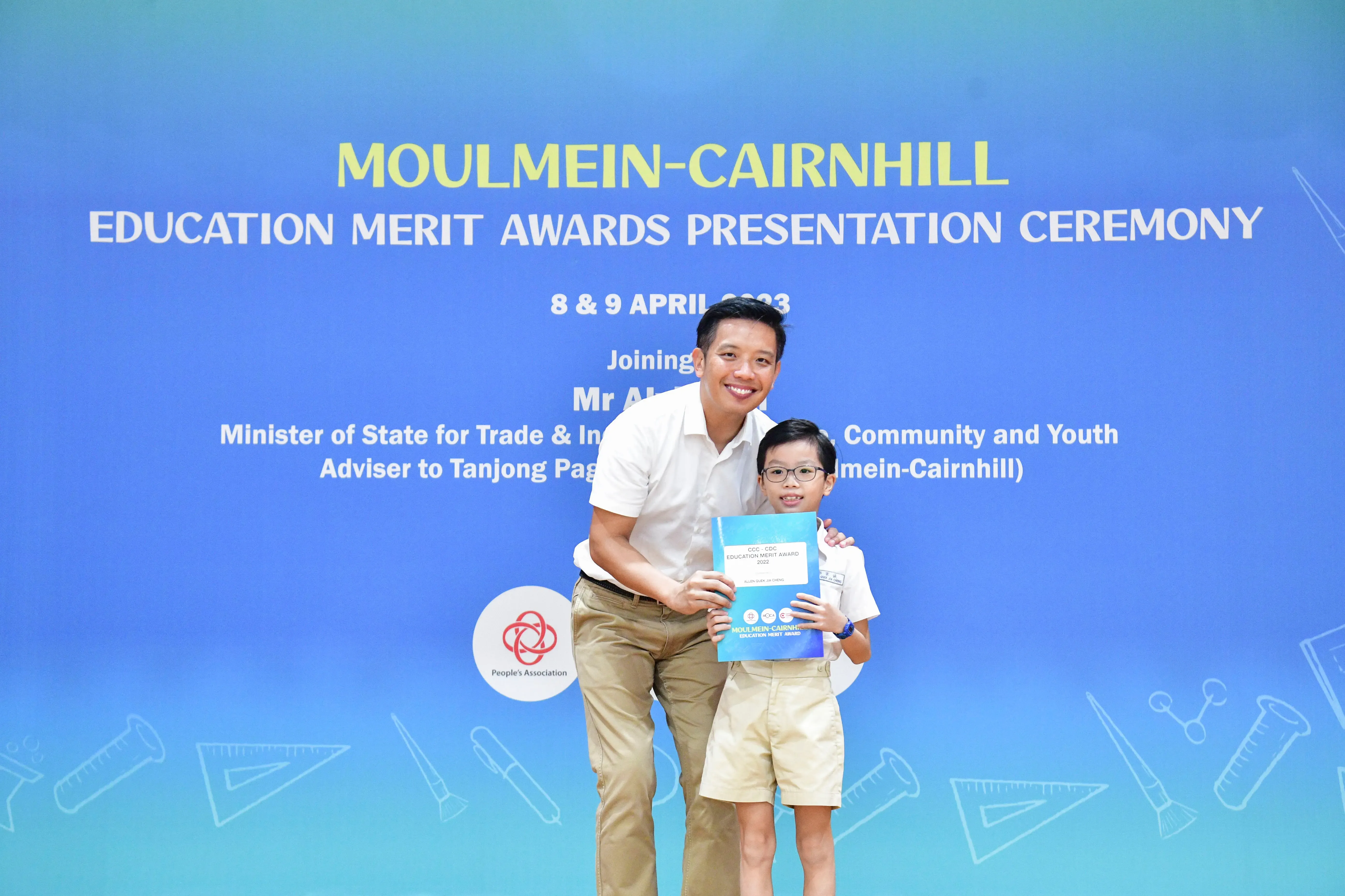 A man and a young boy holding a blue award folder stand before a Moulmein-Cairnhill Education Merit Awards banner.