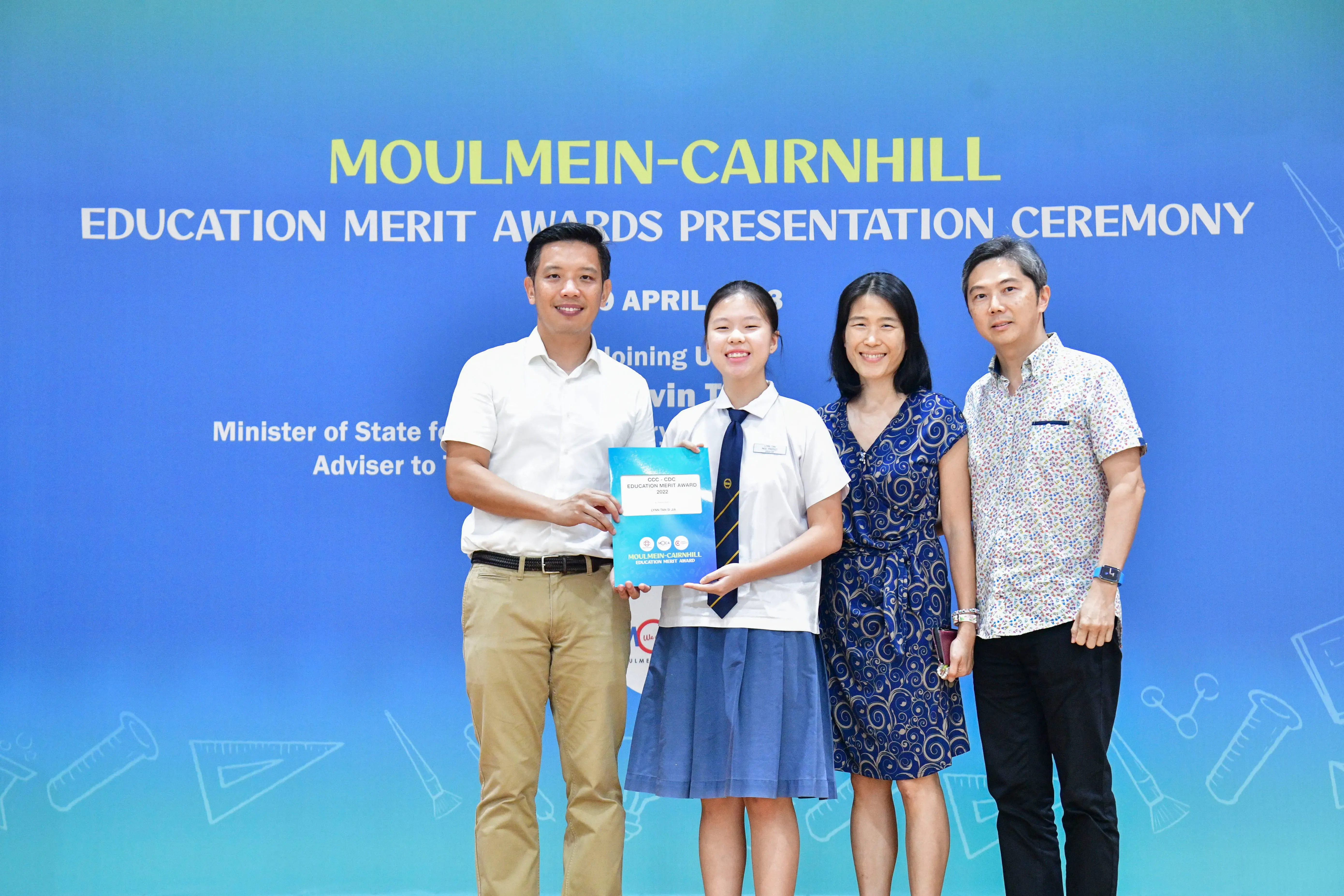 Four people pose at the Moulmein-Cairnhill Education Merit Awards Presentation Ceremony, with a student holding an award folder.