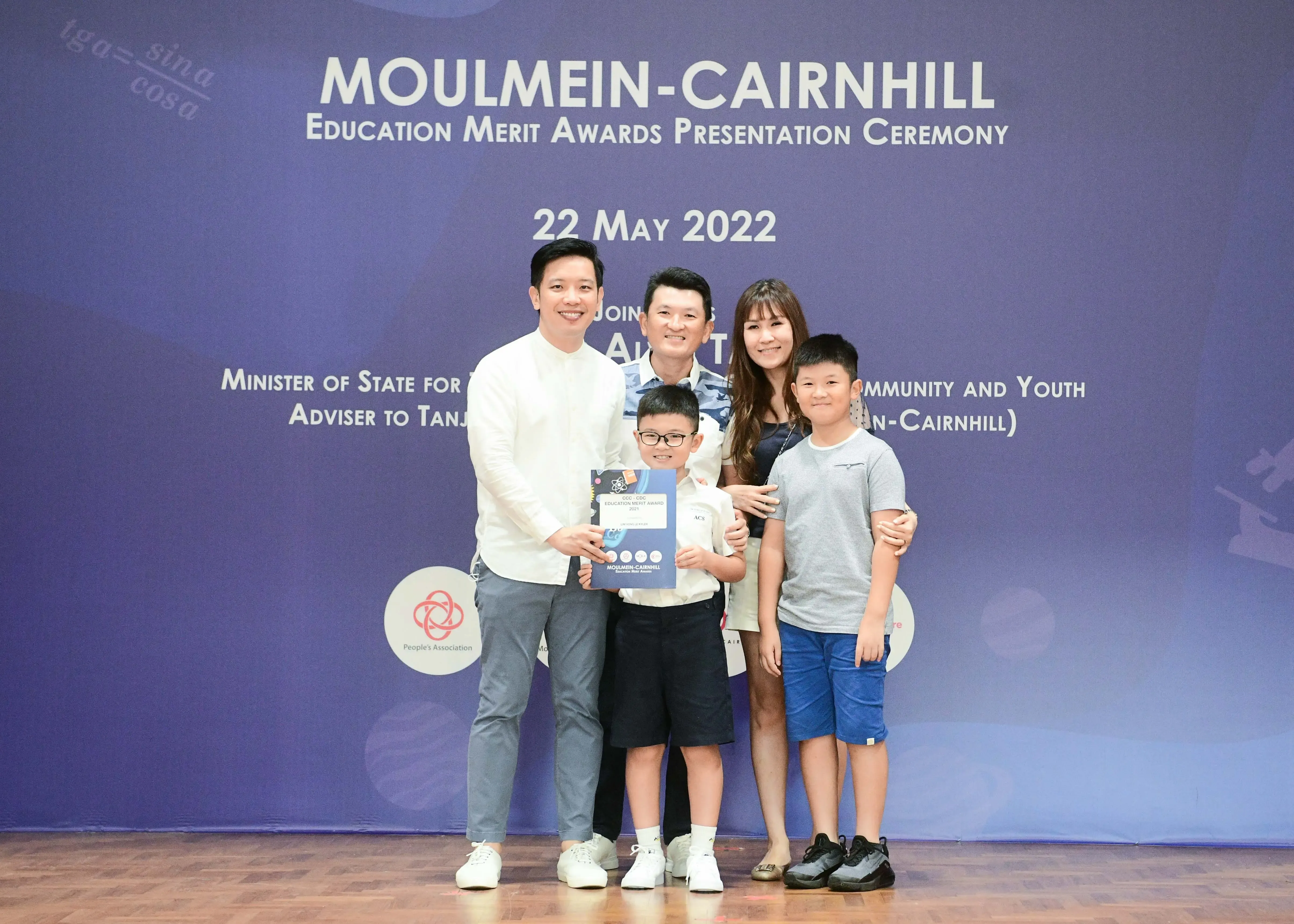 Five people pose in front of a Moulmein-Cairnhill Education Merit Awards banner dated 22 May 2022.
