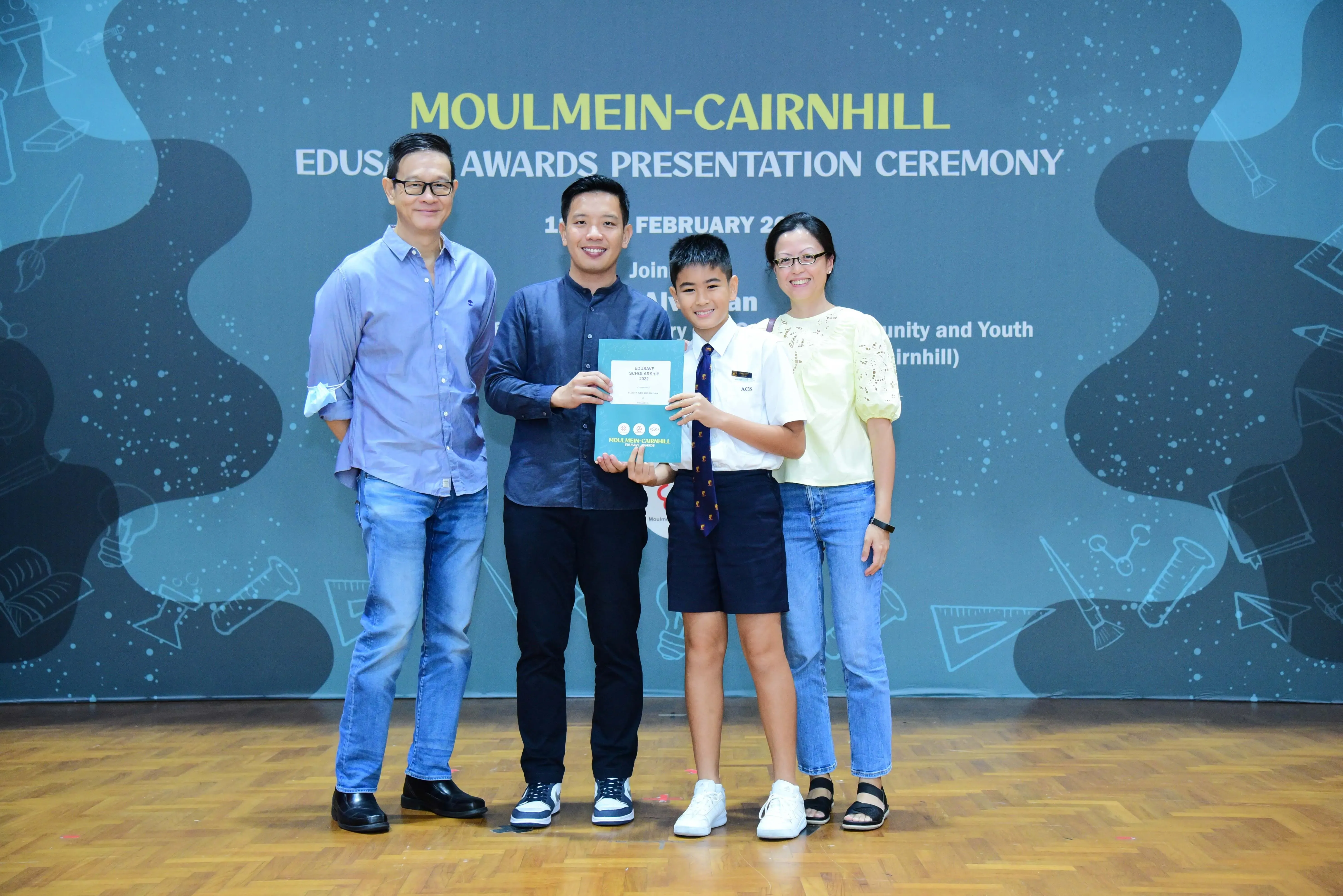 Four people pose at the Moulmein-Cairnhill Edusave Awards Presentation Ceremony, with a boy holding an award certificate.