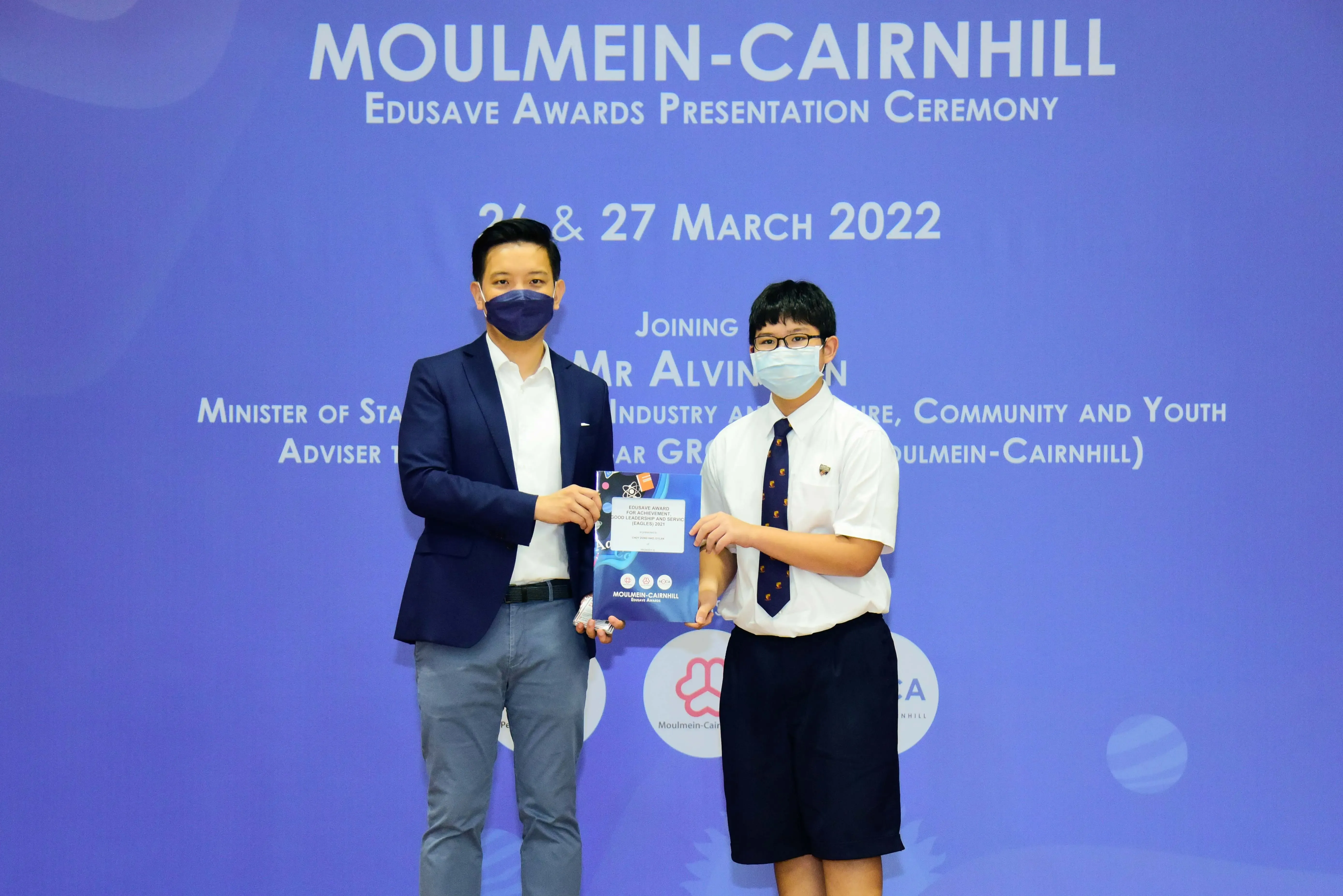 Two masked individuals hold an award booklet at the Moulmein-Cairnhill Edusave Awards Presentation Ceremony, March 2022.
