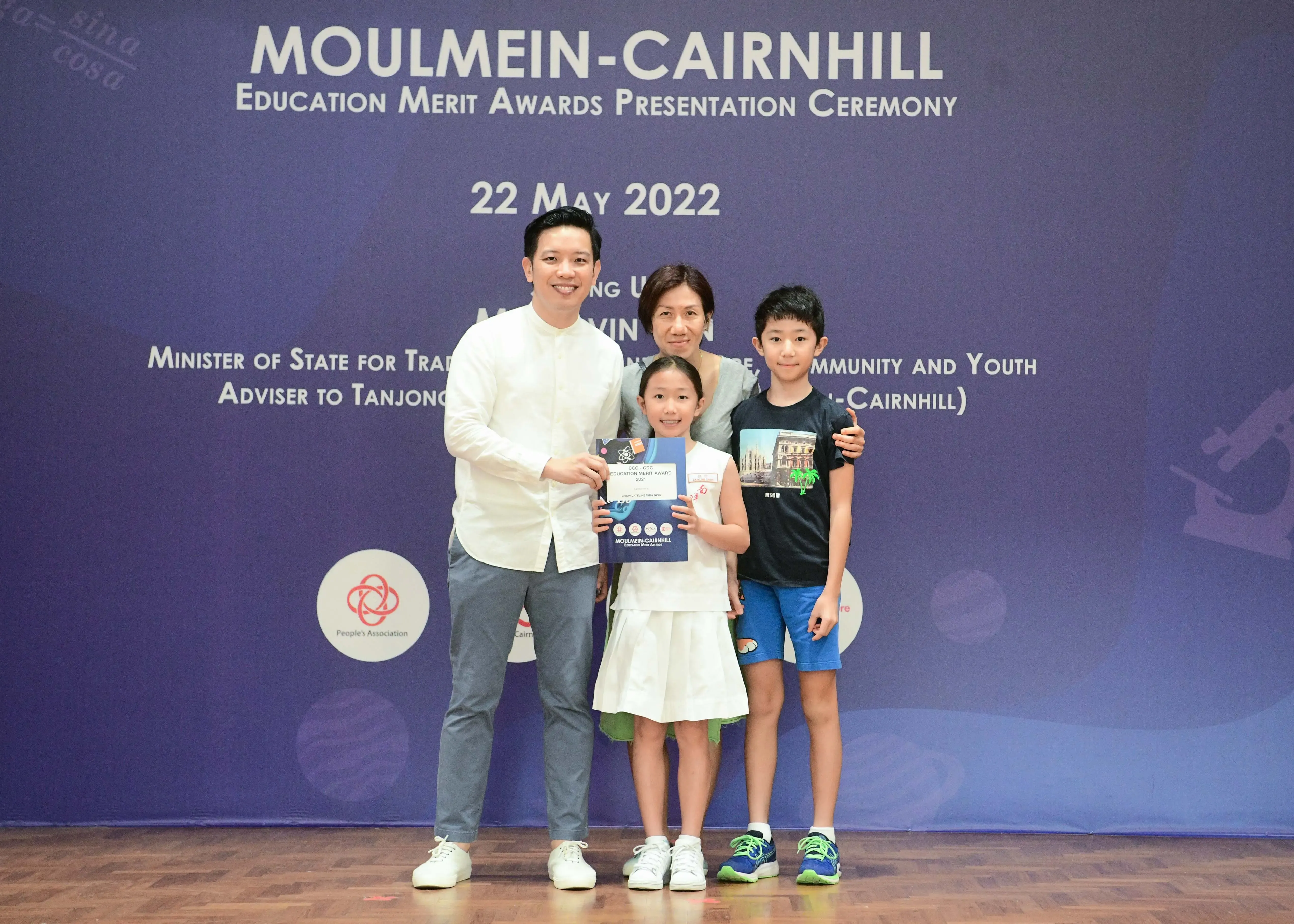Four people pose in front of a Moulmein-Cairnhill Education Merit Awards banner dated 22 May 2022.
