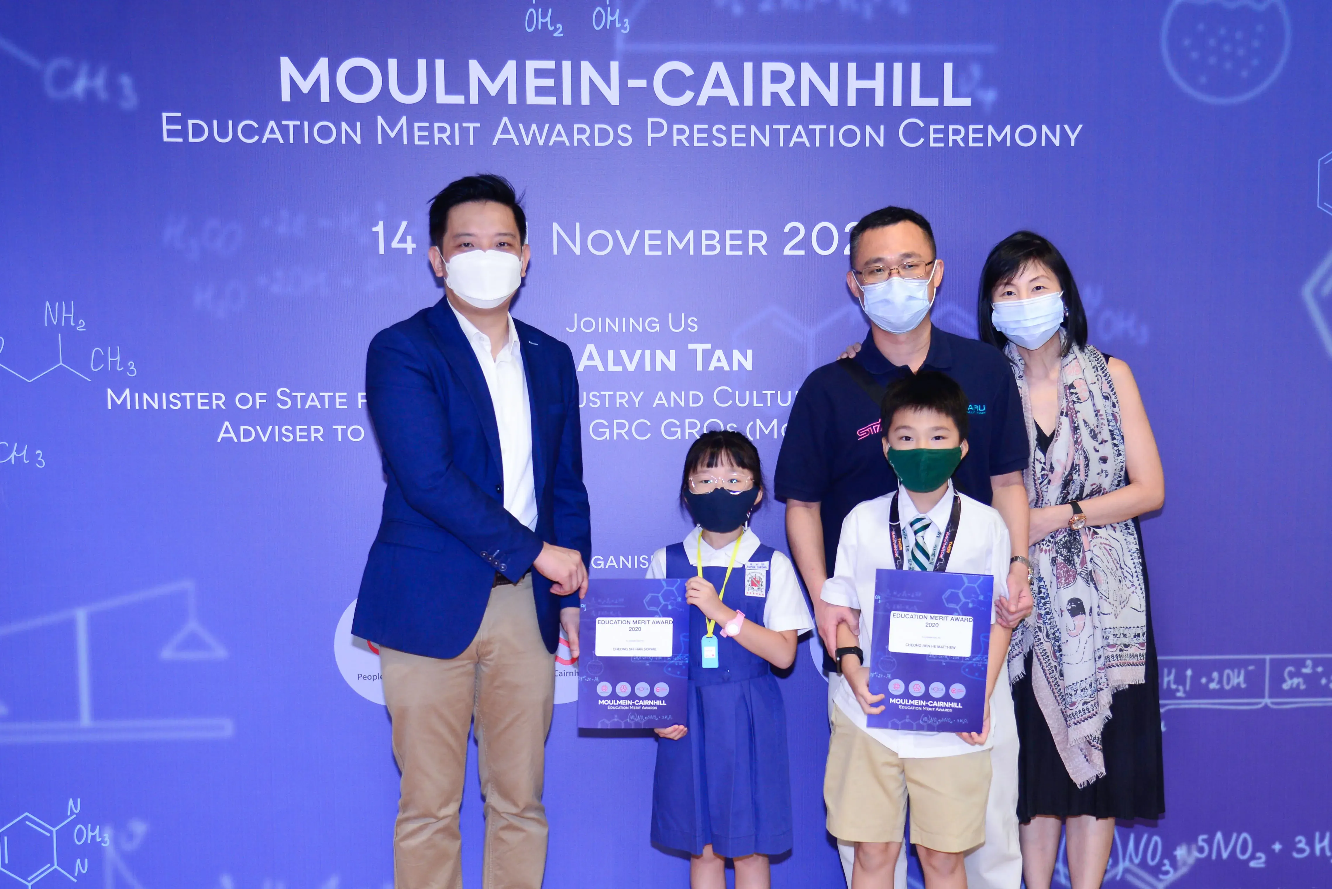 Five masked people pose at the Moulmein-Cairnhill Education Merit Awards Ceremony, two children holding award certificates.
