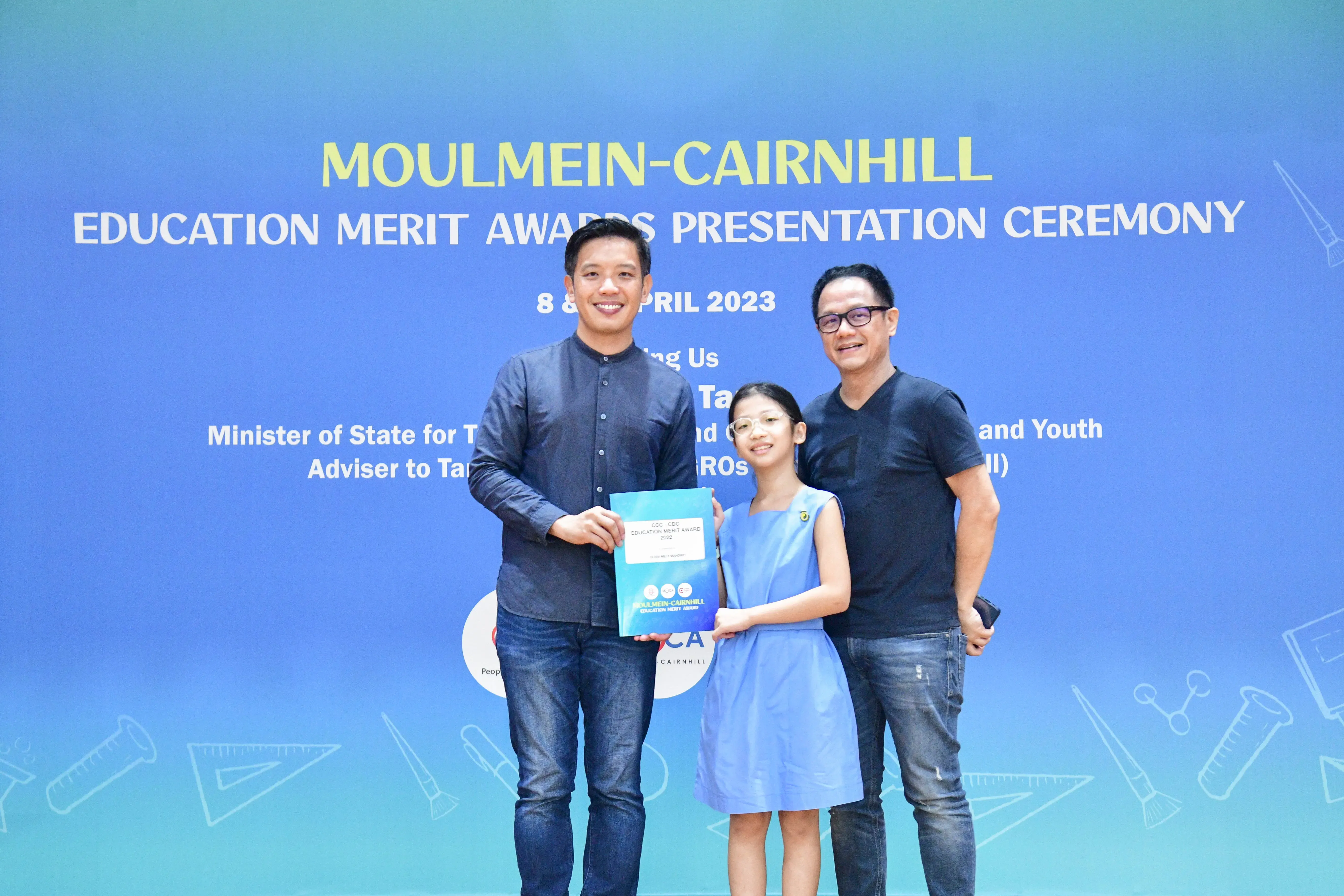 Two men and a girl holding a certificate pose before the Moulmein-Cairnhill Education Merit Awards 2023 banner.