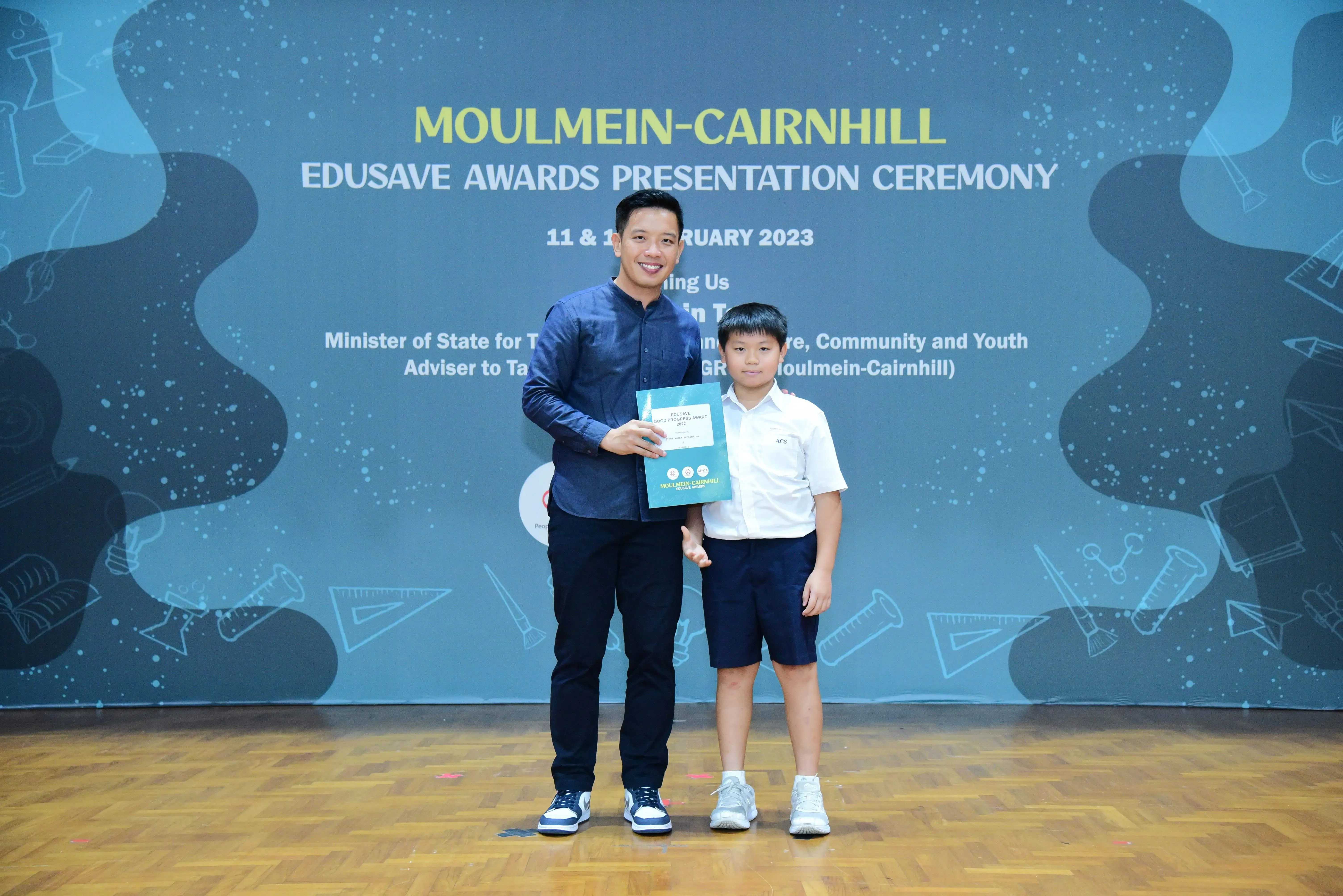 An adult and a young boy pose together holding a certificate at the Moulmein-Cairnhill Edusave Awards Ceremony 2023.