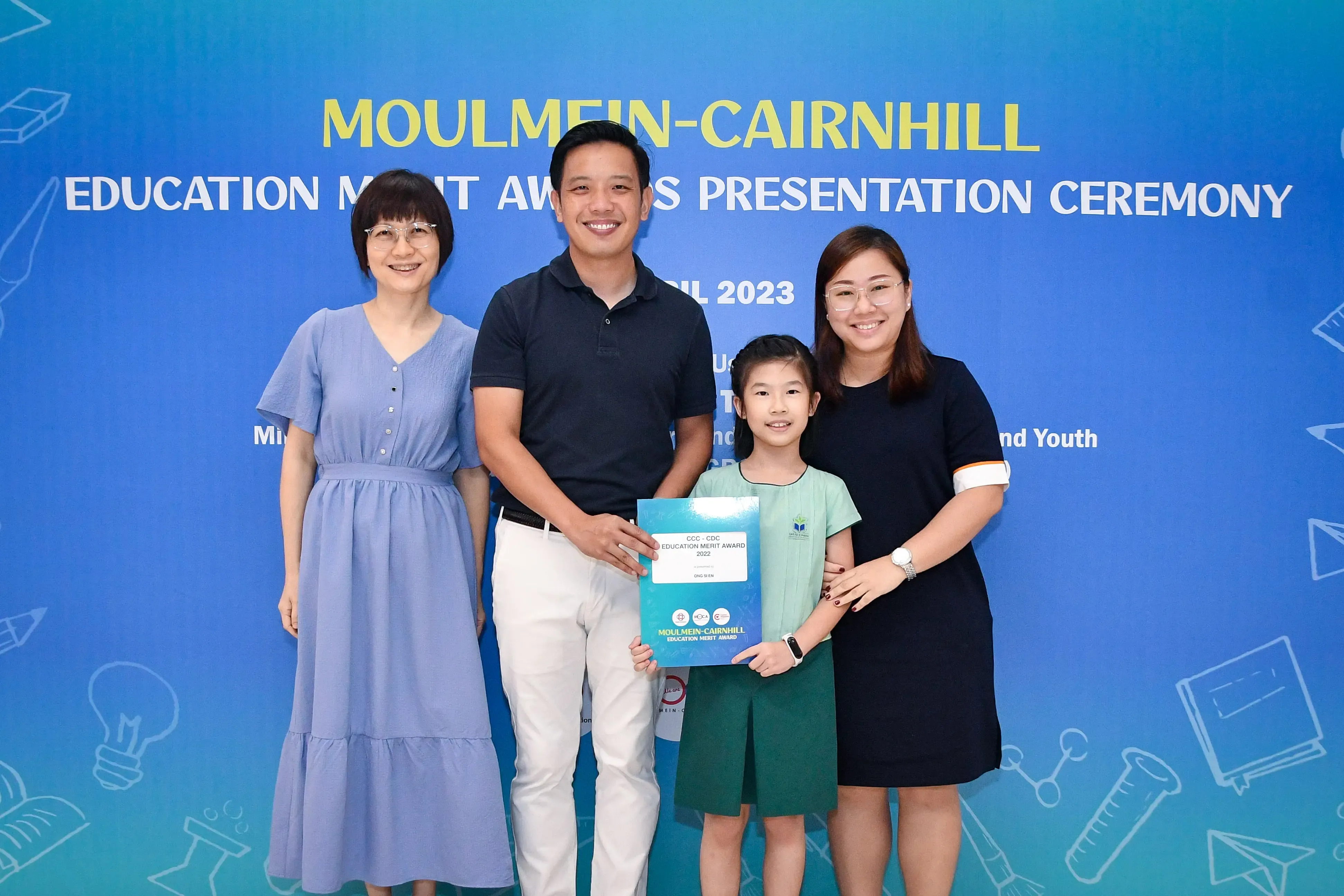 Three adults and a young girl pose at the 2023 Moulmein-Cairnhill Education Merit Awards ceremony, holding a certificate.