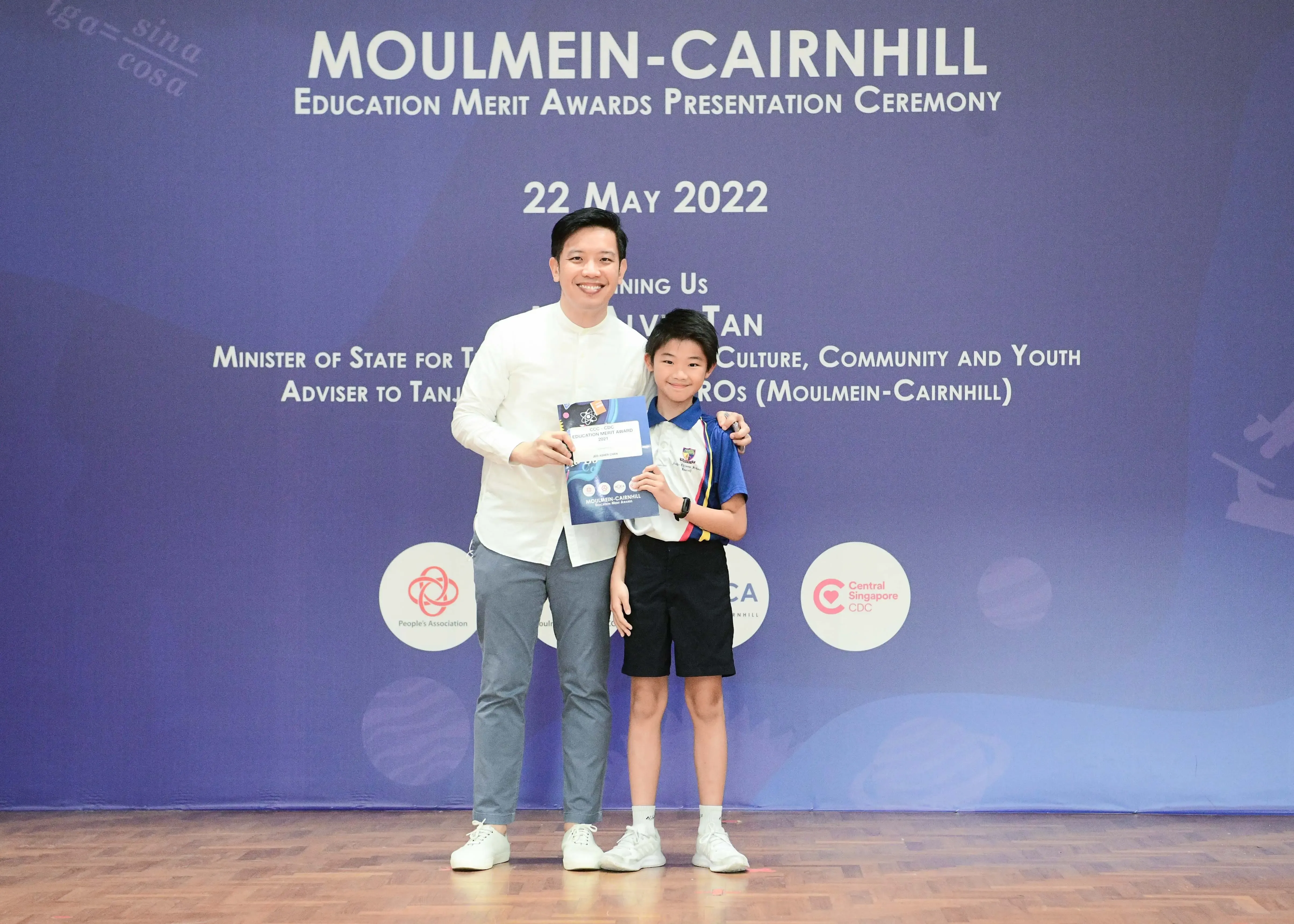 An adult and a young student pose holding a certificate at the Moulmein-Cairnhill Education Merit Awards ceremony on 22 May 2022.