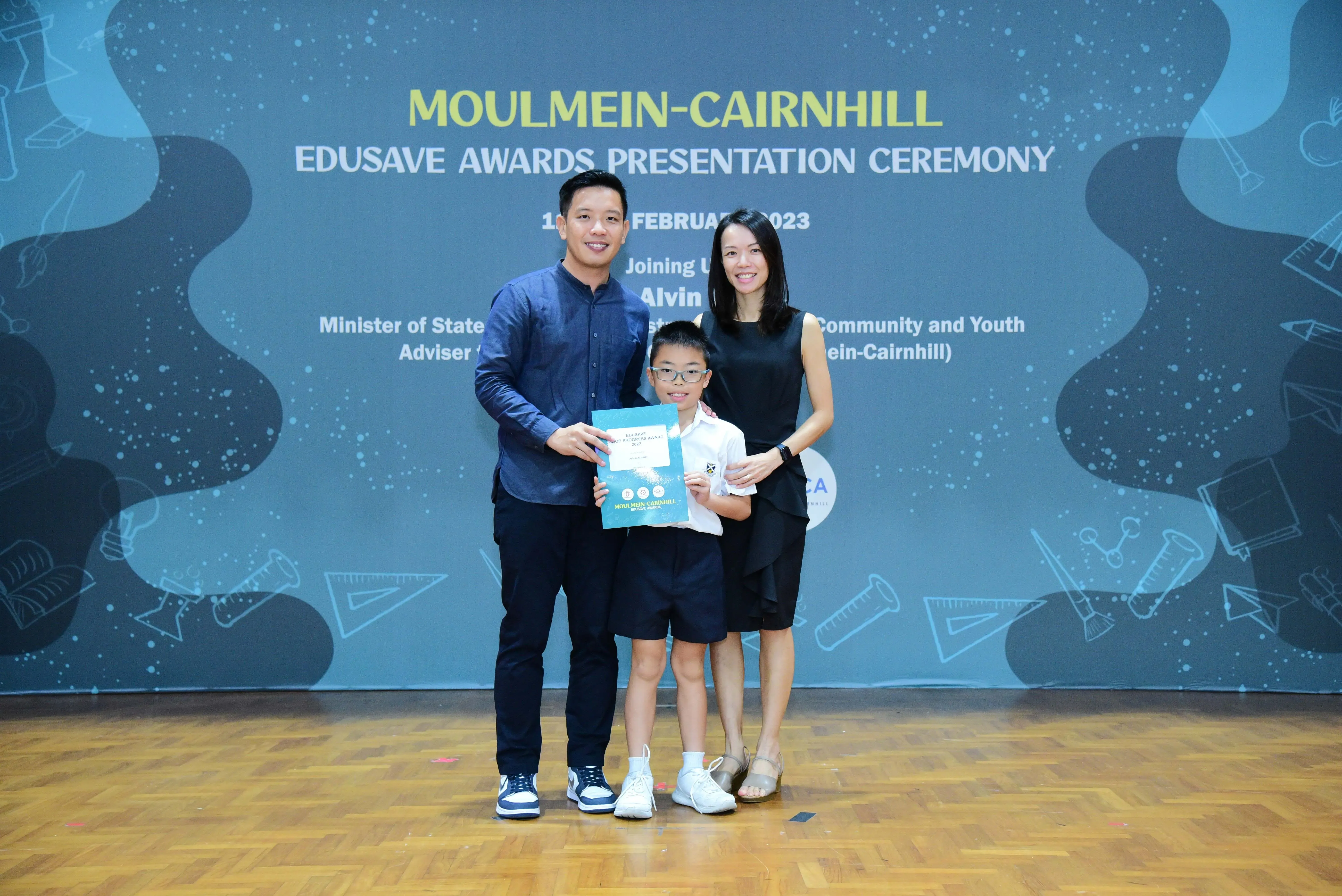 Two adults and a young boy holding a certificate pose at the Moulmein-Cairnhill Edusave Awards Ceremony 2023.