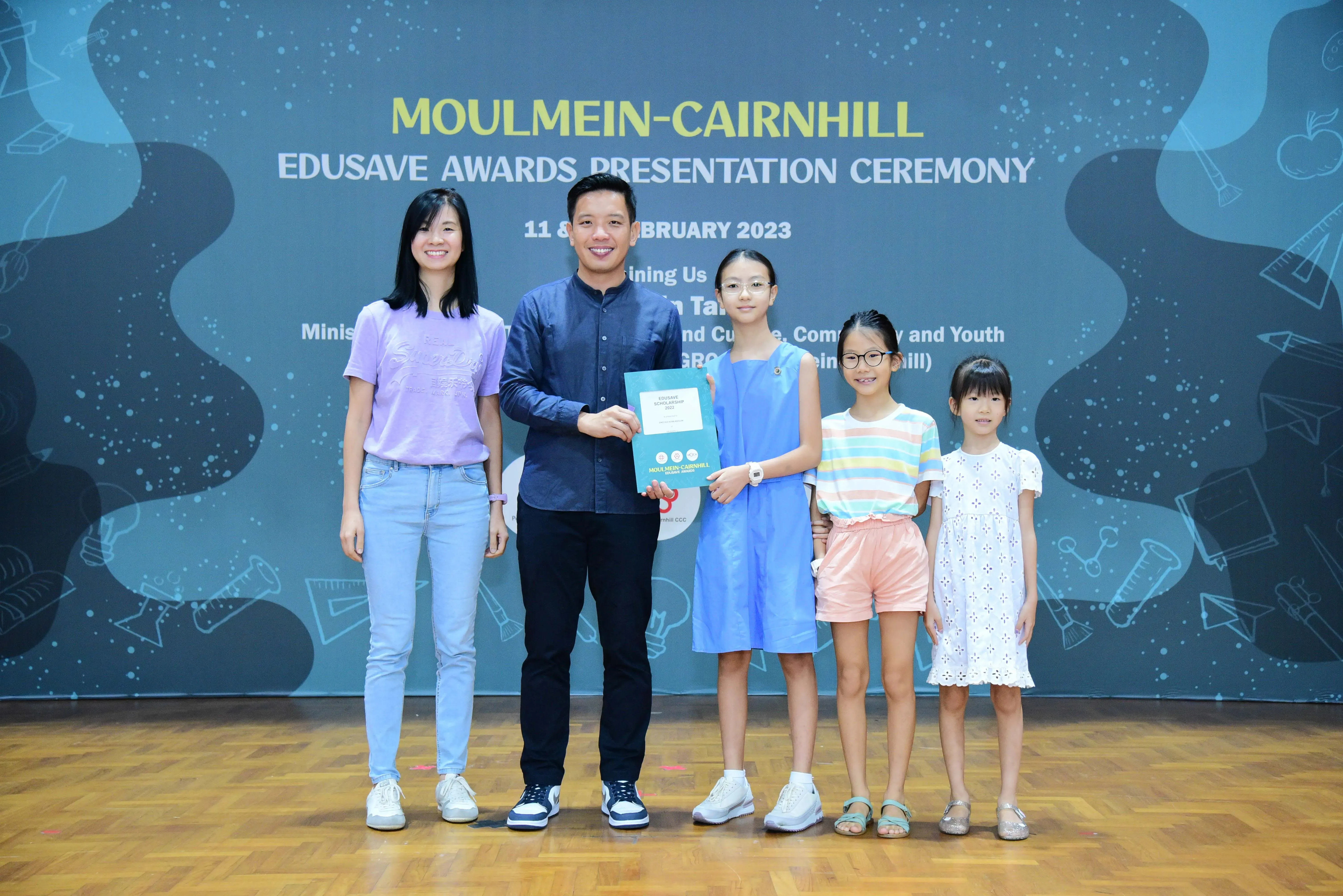 A man presents an award to a girl among three children and a woman at the Moulmein-Cairnhill Edusave Awards Ceremony 2023.
