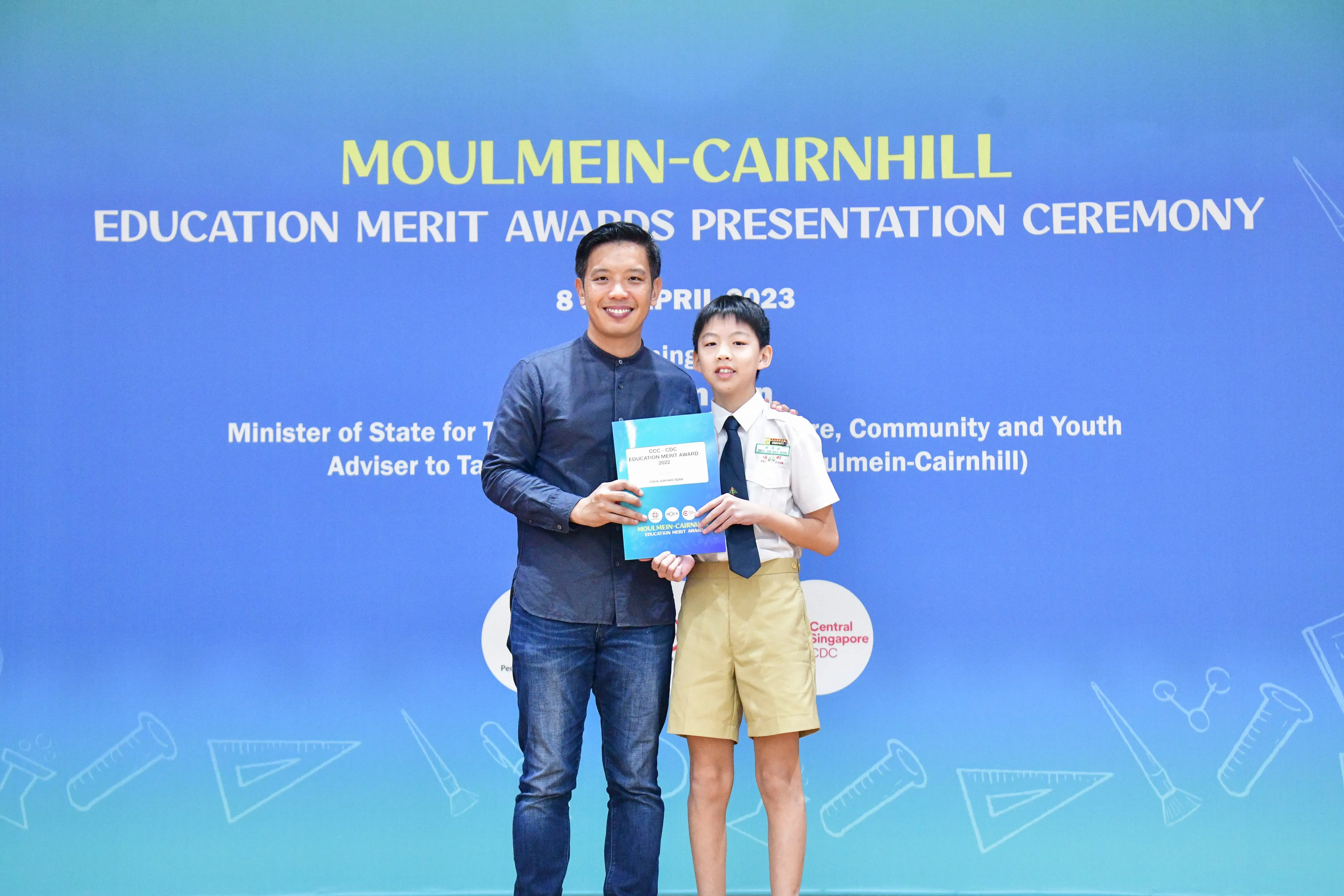 An adult and a uniformed student hold an award together at the Moulmein-Cairnhill Education Merit Awards Ceremony 2023.