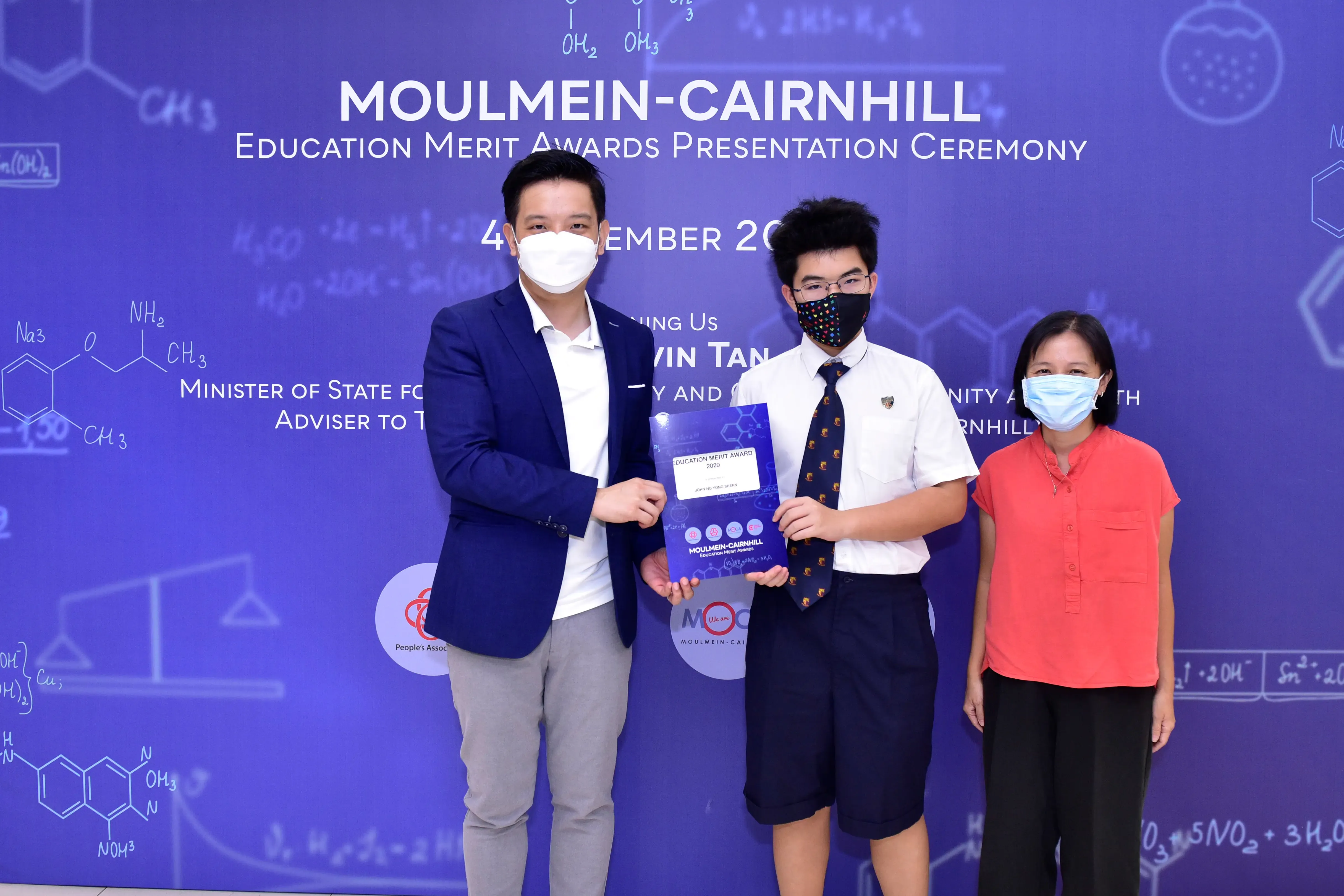 Three masked people pose at the Moulmein-Cairnhill Education Merit Awards Presentation Ceremony, holding an award booklet.