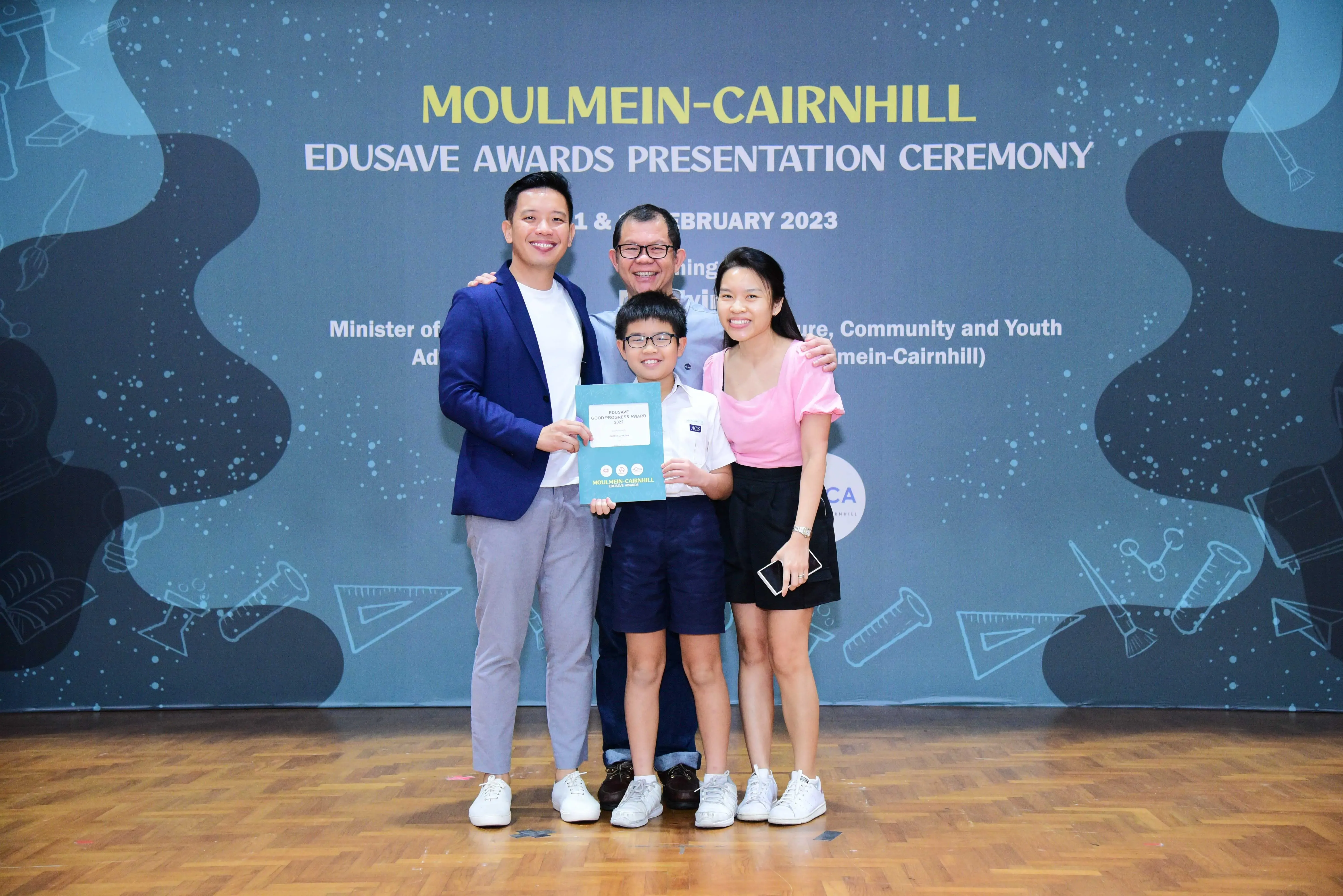 Four people smile together at the Moulmein-Cairnhill Edusave Awards Presentation Ceremony, with a boy holding a certificate.