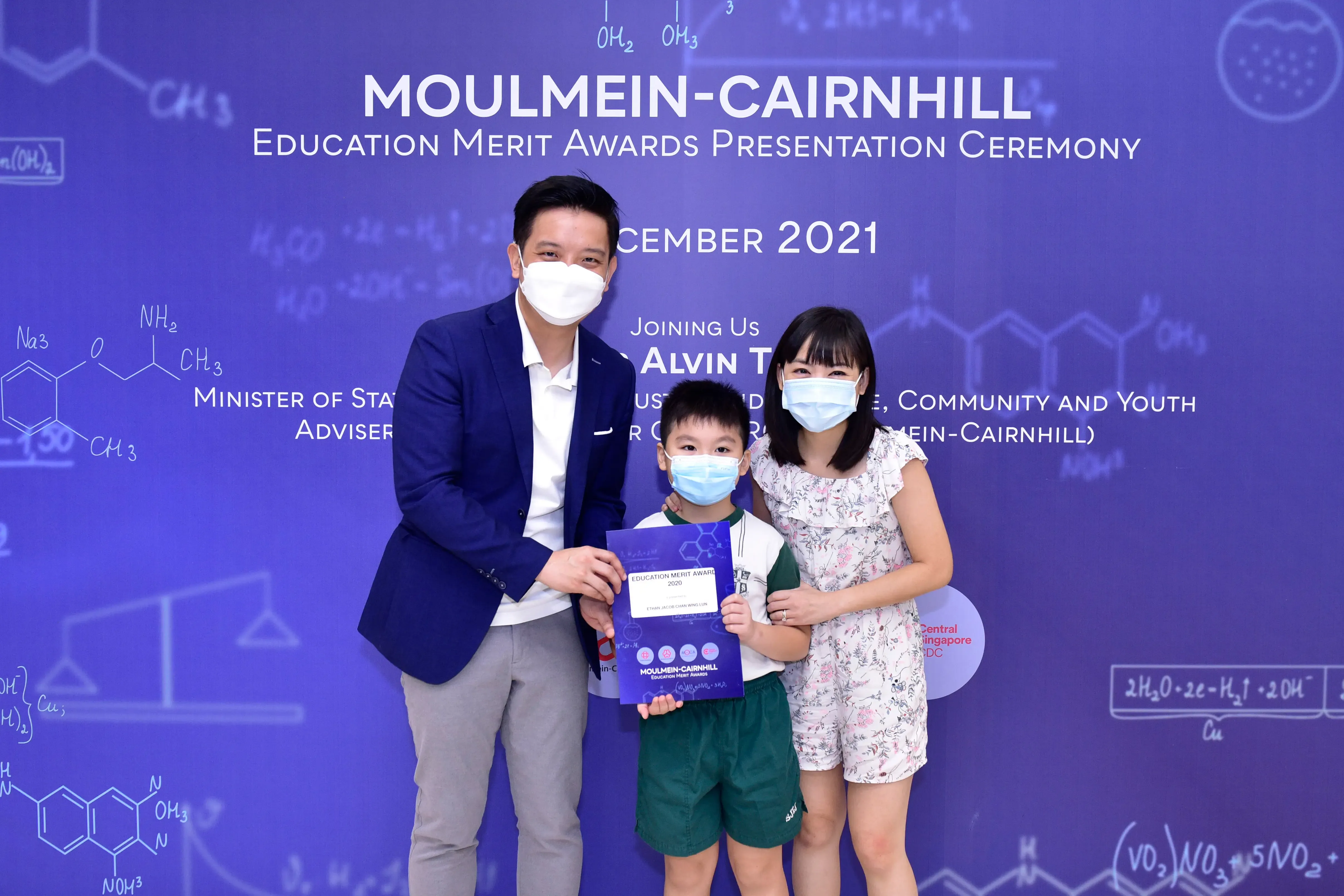 Two adults and a child wearing masks hold a certificate at the Moulmein-Cairnhill Education Merit Awards 2021.