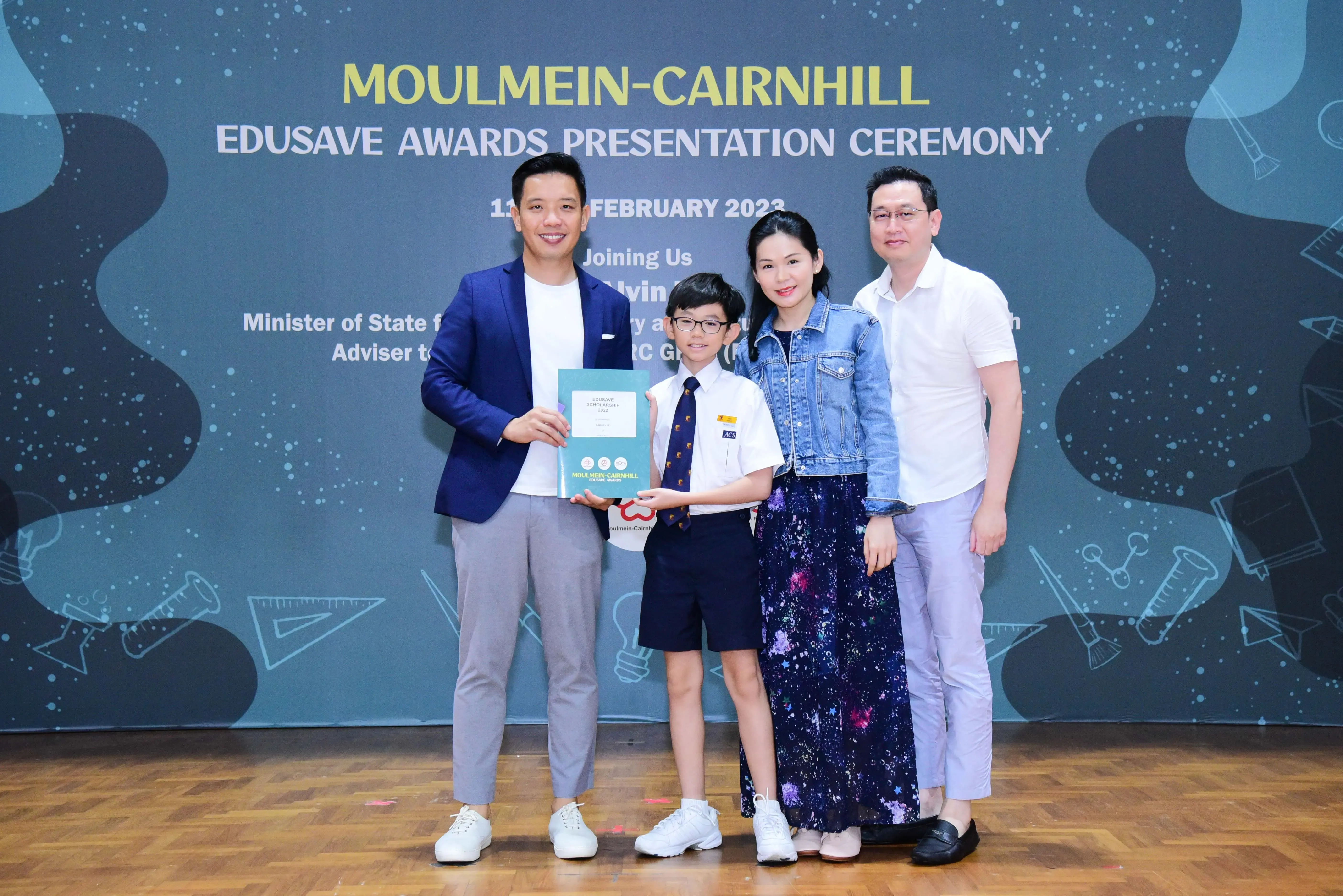 Four people pose at the Moulmein-Cairnhill Edusave Awards Ceremony, with a boy in uniform holding a certificate.