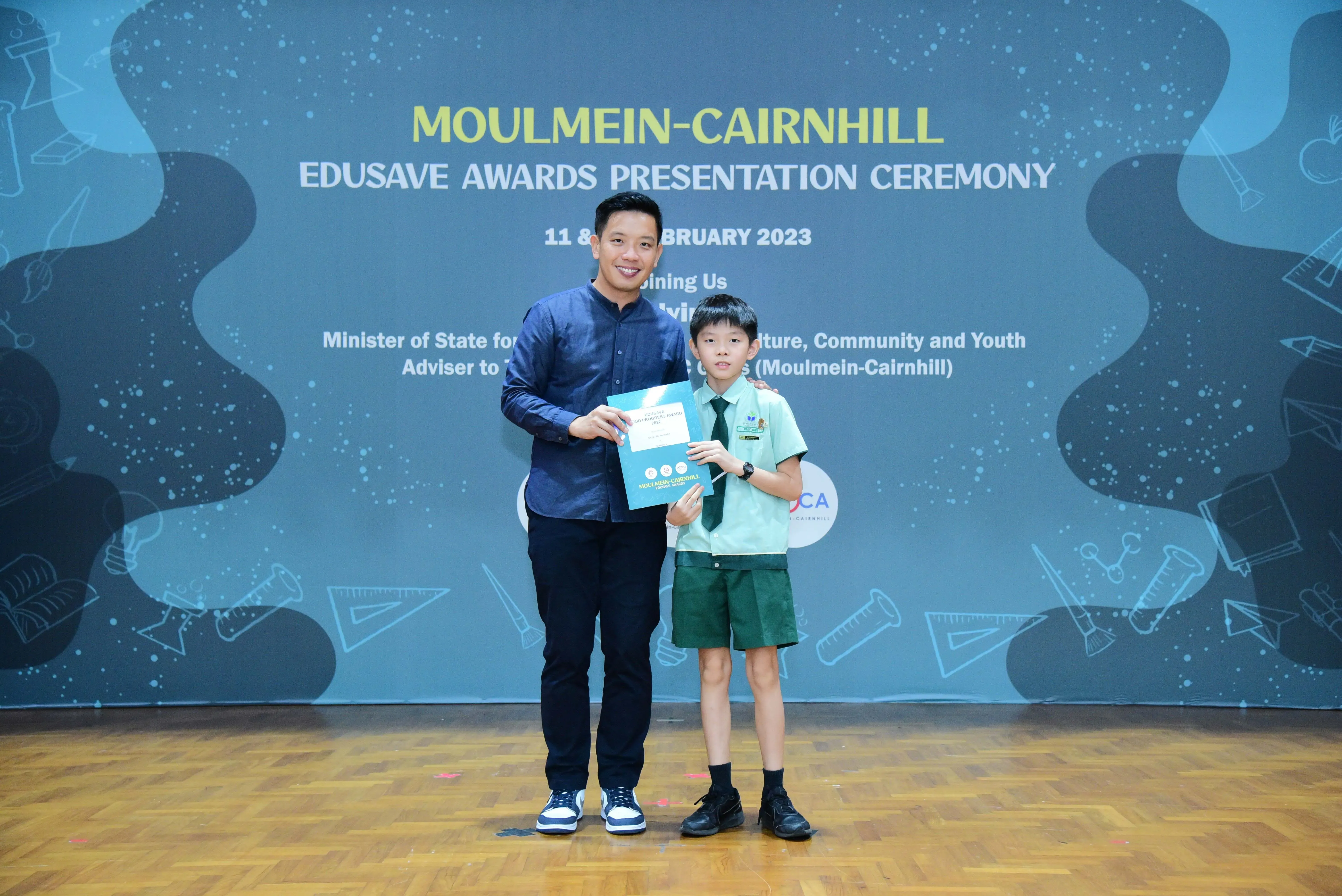 A man and a young boy in school uniform hold a certificate at the Moulmein-Cairnhill Edusave Awards Ceremony 2023.